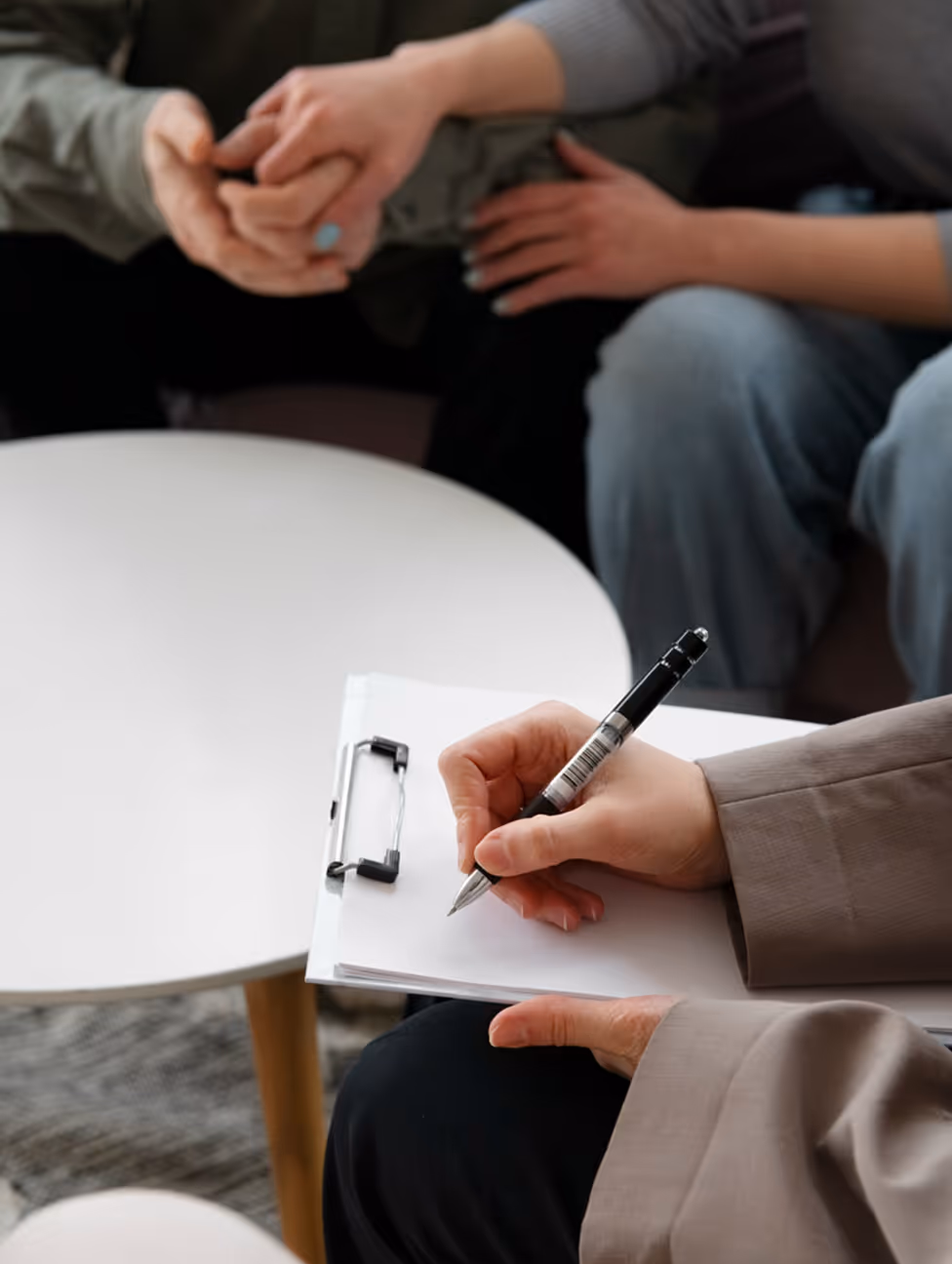 A close-up of a therapist writing notes on a clipboard while two people hold hands in the background.