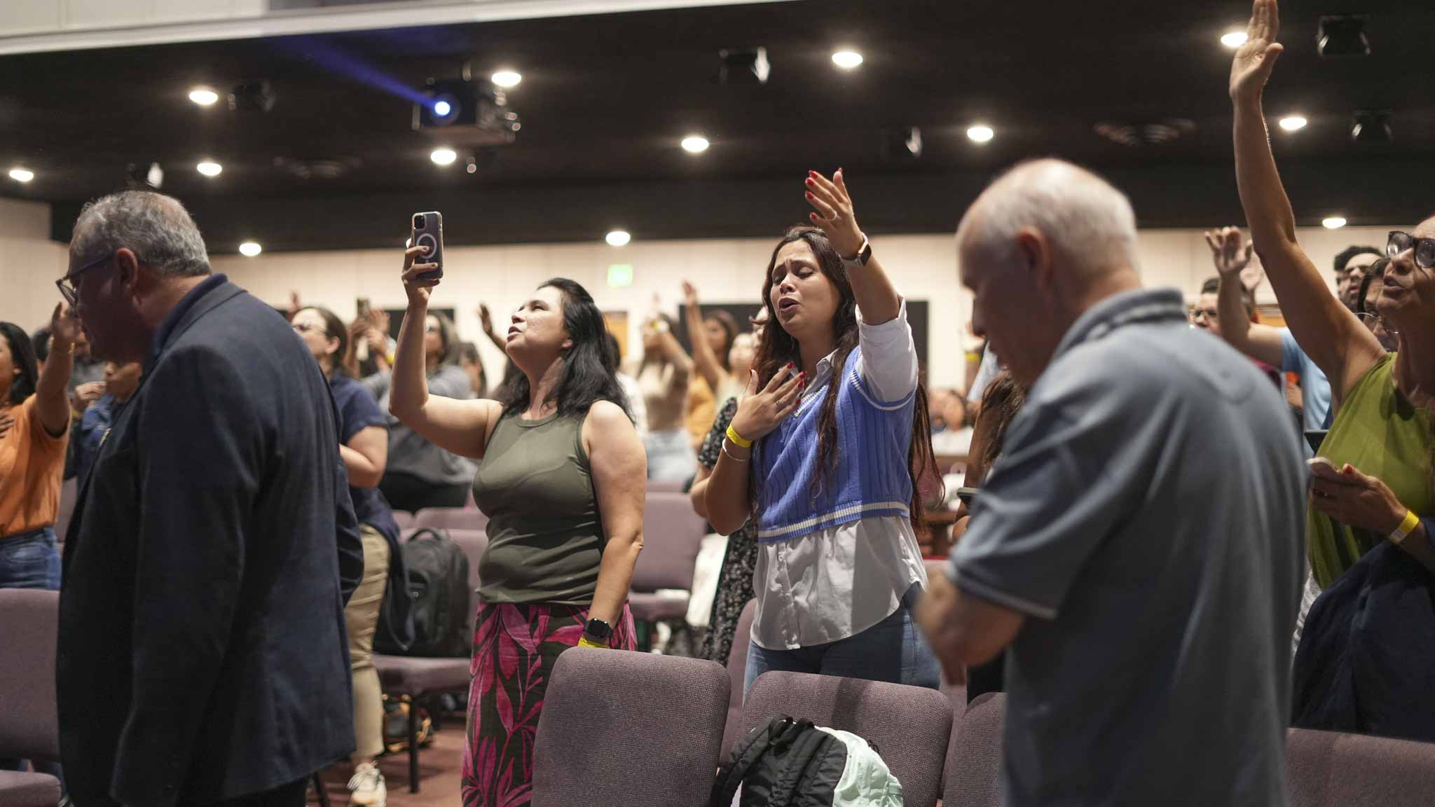 People in an indoor gathering raising hands and singing or praying with eyes closed or looking upward.