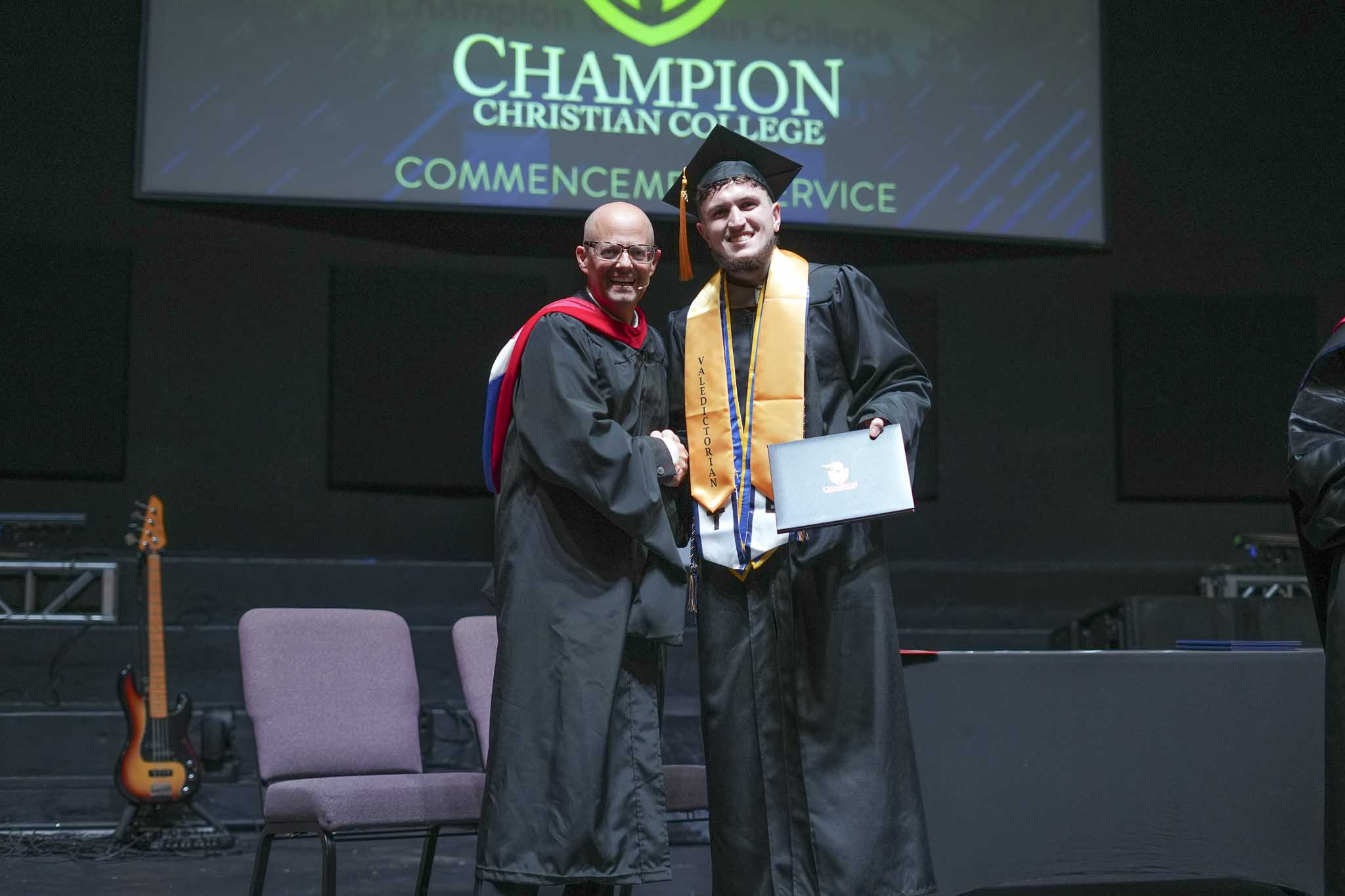 Graduate in cap and gown with valedictorian stole shaking hands with faculty member during Champion Christian College commencement service.