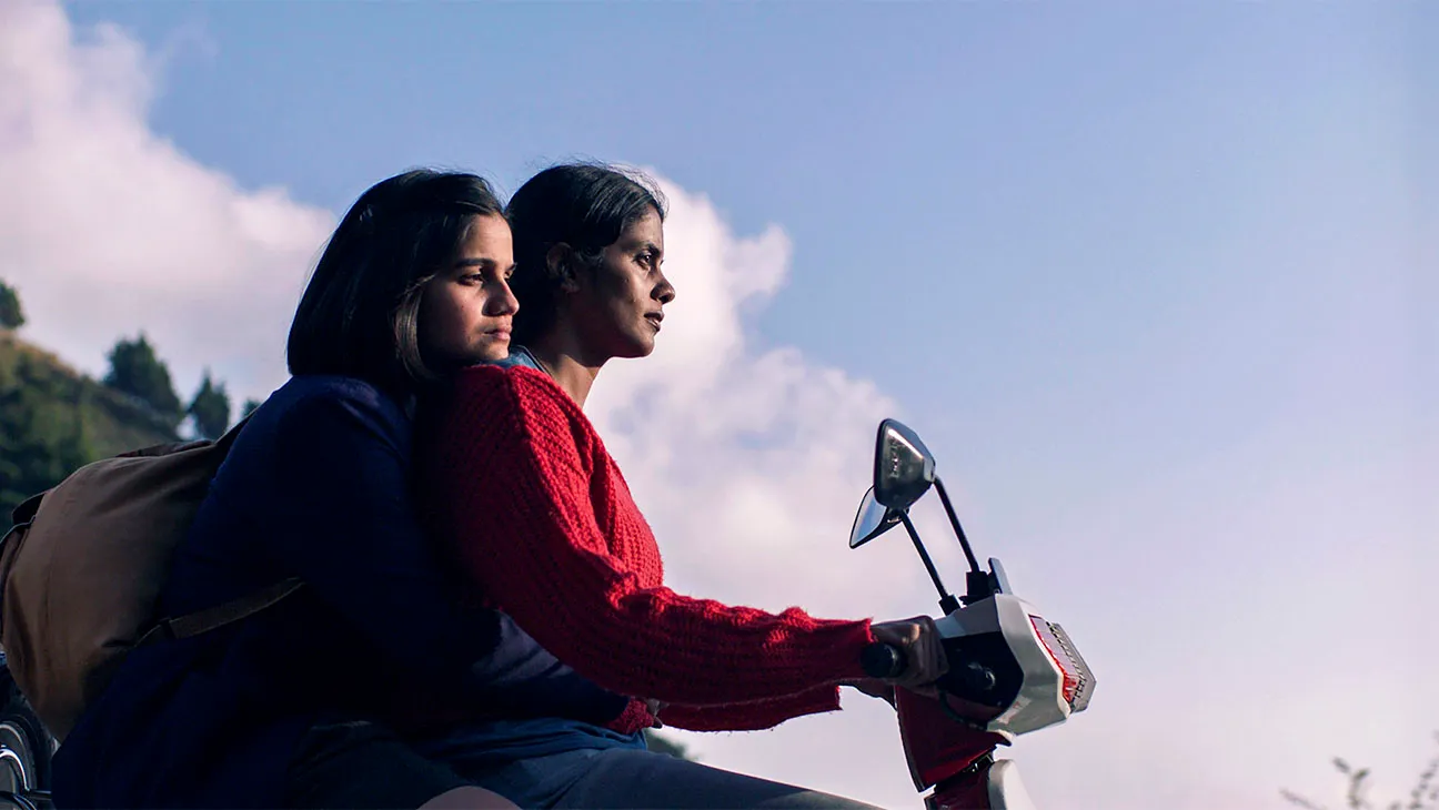 Movie still from Girls Will Be Girls. A young girl rides on the back of a moped driven by an older woman.
