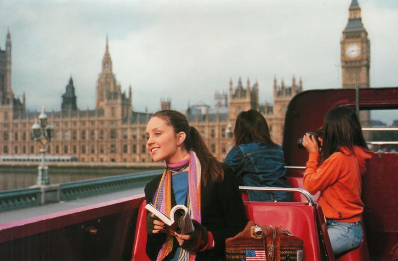 Movie still from What A Girl Wants. A young teen girl smiles with a tour guide book while on a double decker bridge in London, Big Ben in the background.