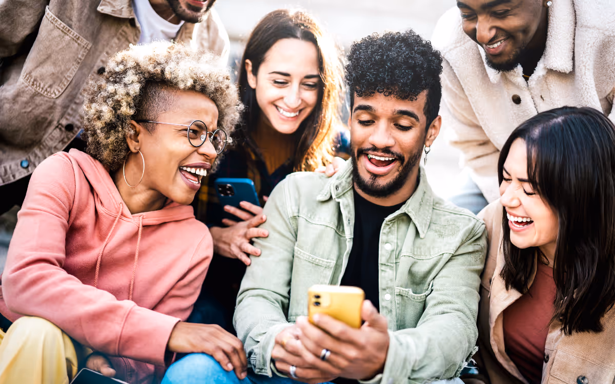 A group of young people crowd around a mobile phone, smiling. 