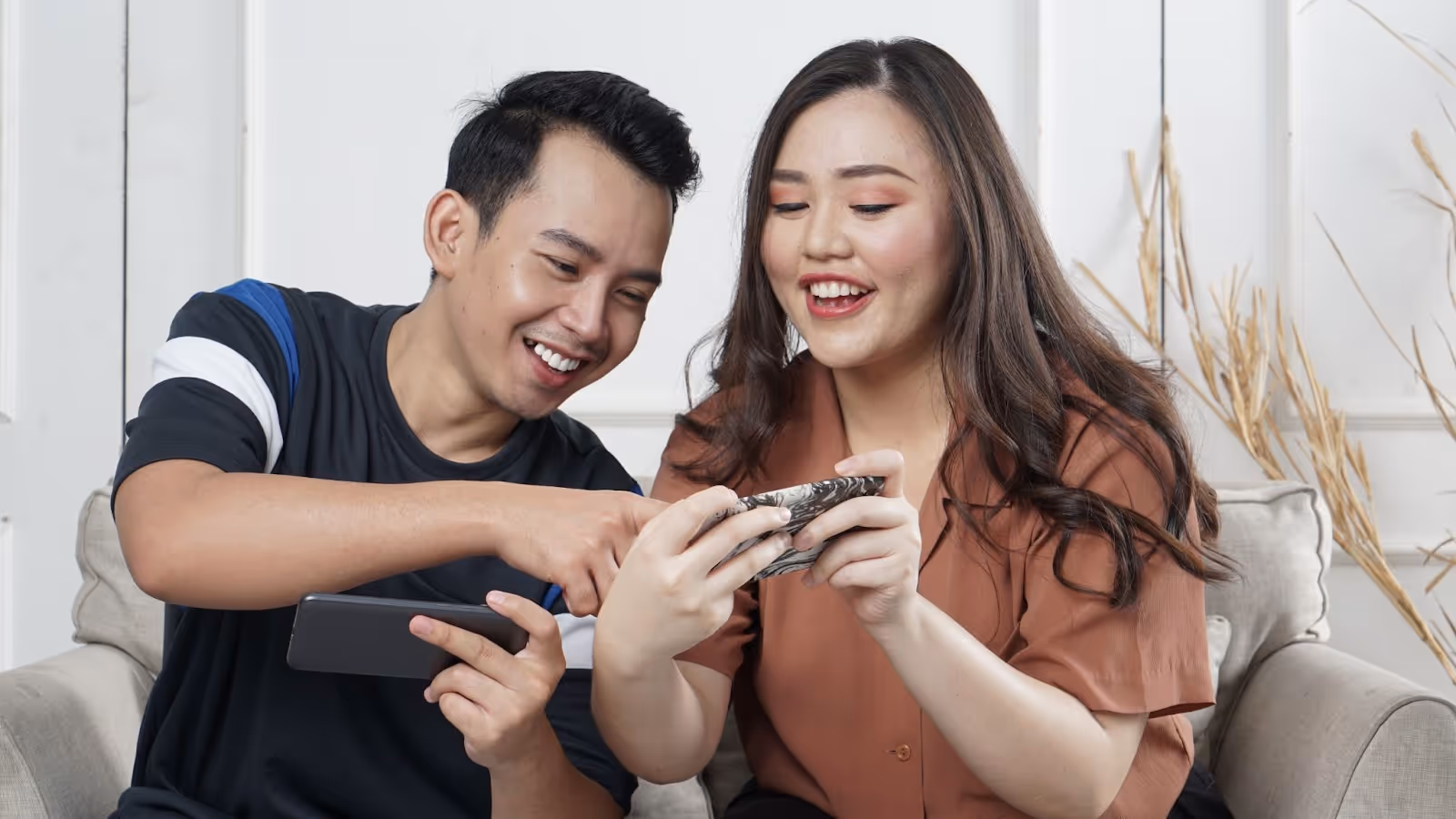 Two people smiling at their mobile phones whilst sitting on a couch.