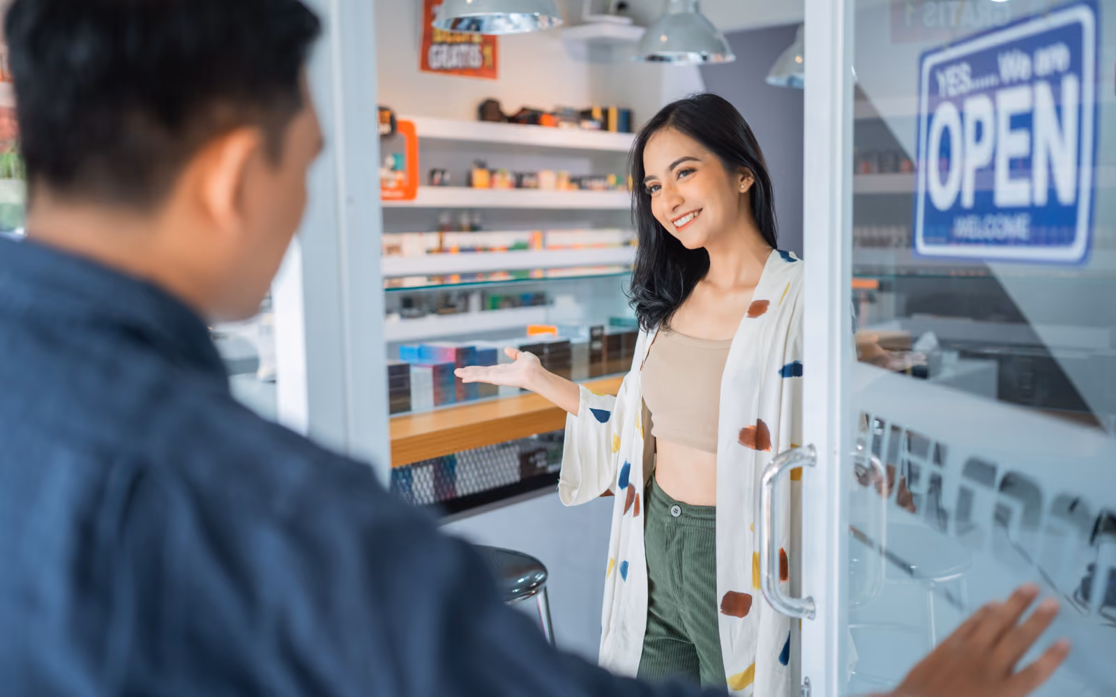 A woman opening the door to a vape store smiling at a customer. 