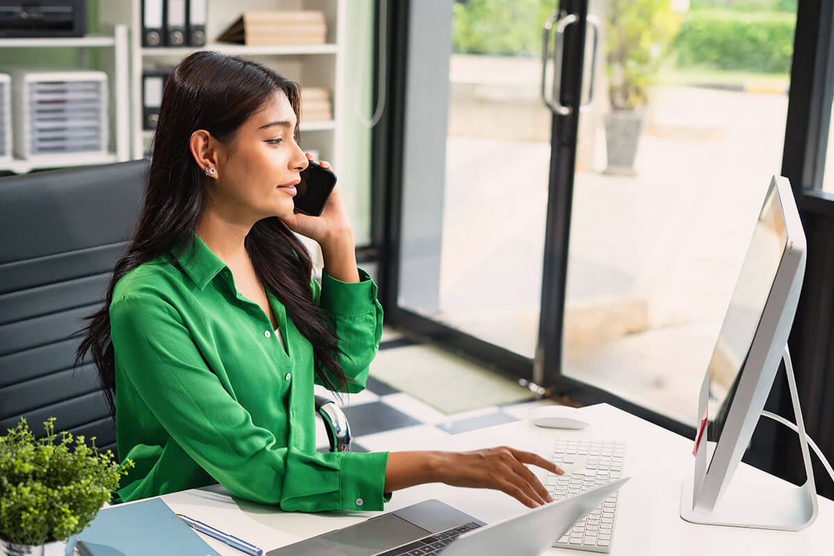 Woman in green shirt talking on phone while typing on a keyboard at a desk with a computer and laptop in a bright office.
