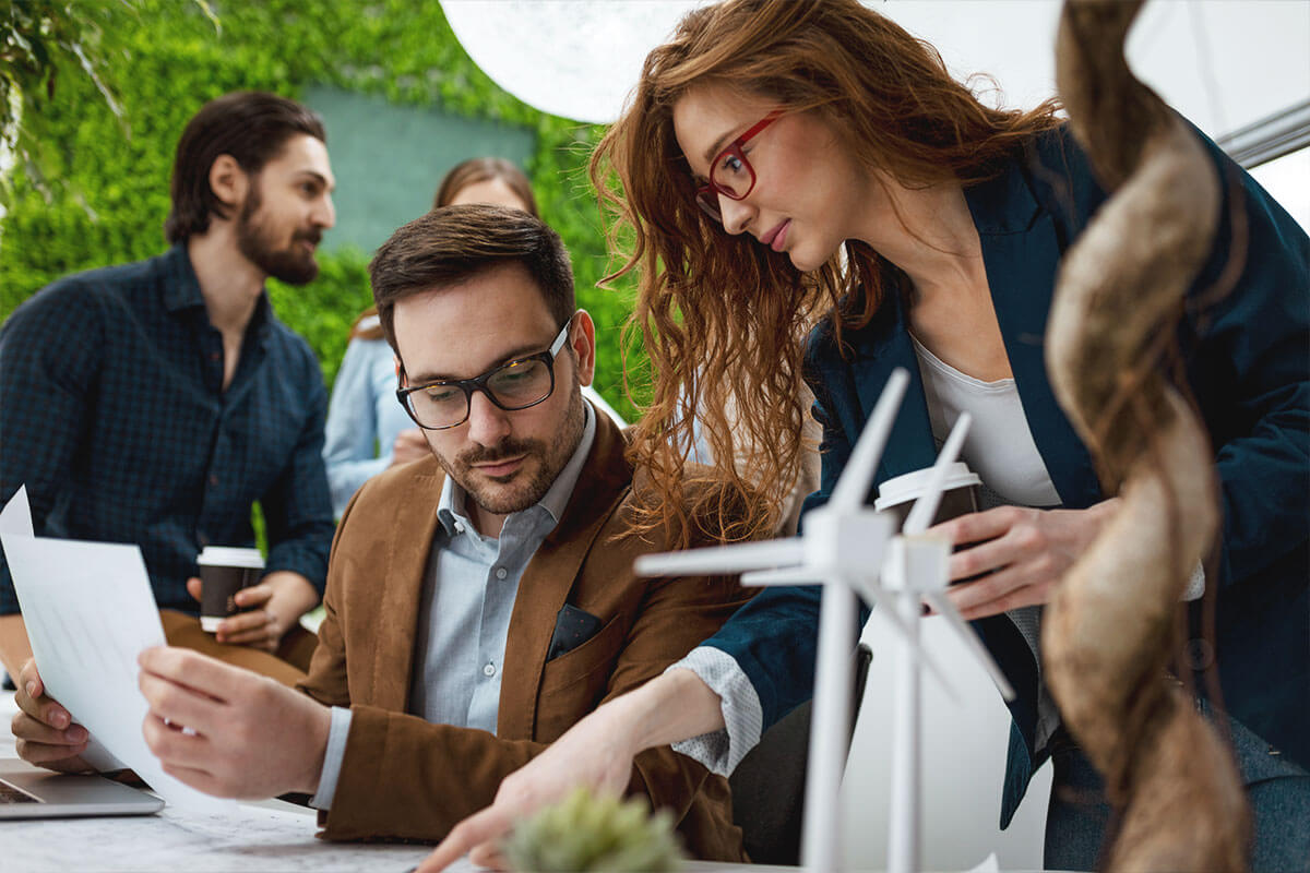 Two professionals discussing documents at a desk with small wind turbine models, with two colleagues in the background.
