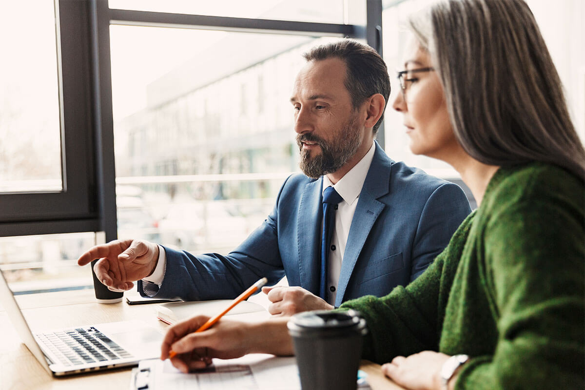 Man in blue suit pointing at a laptop screen while sitting next to a woman in a green sweater who is writing with a pencil at a desk by a window.