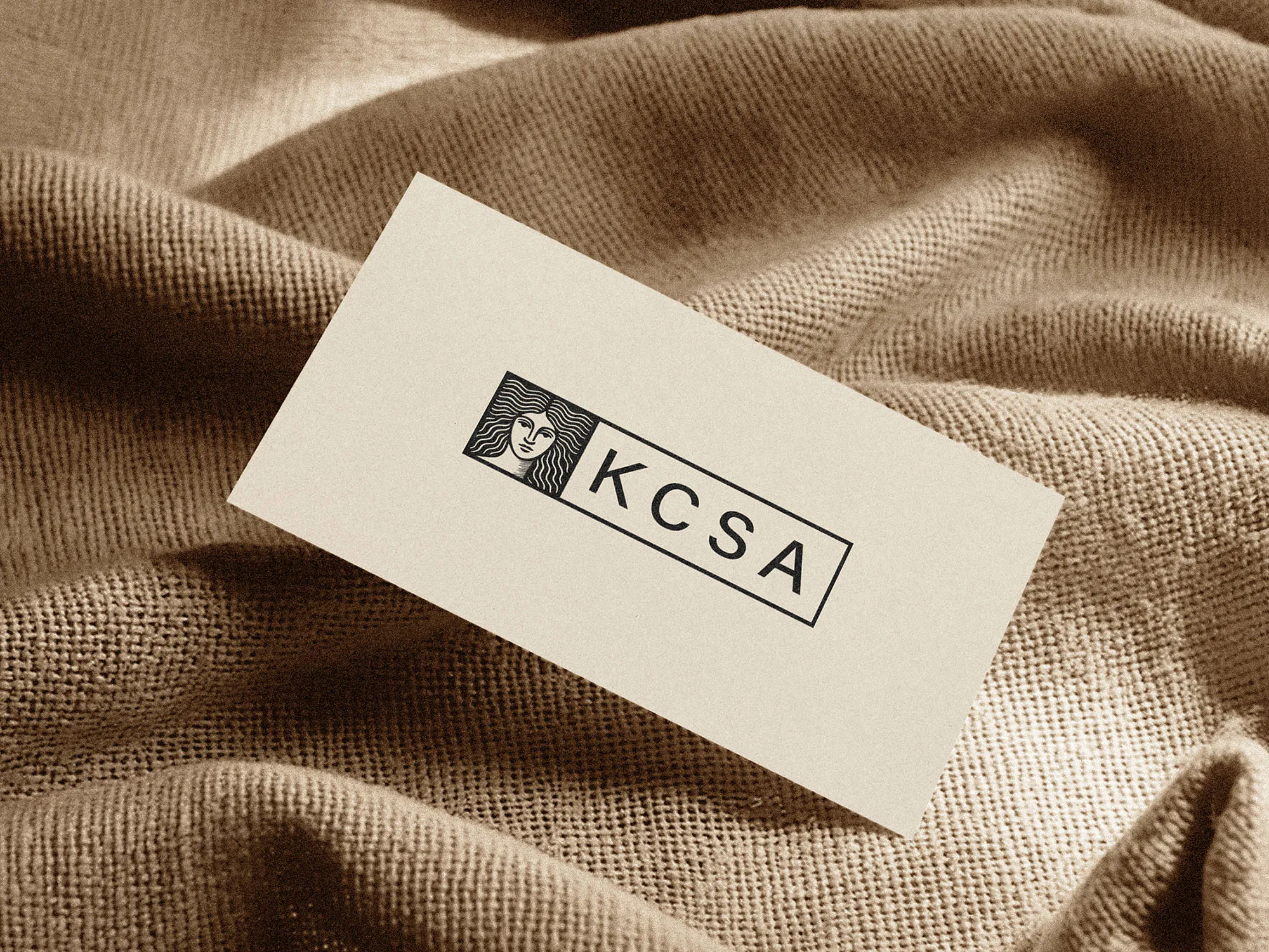 Kate Casprowiak Scher Art logo printed on a cream business card resting on beige fabric.