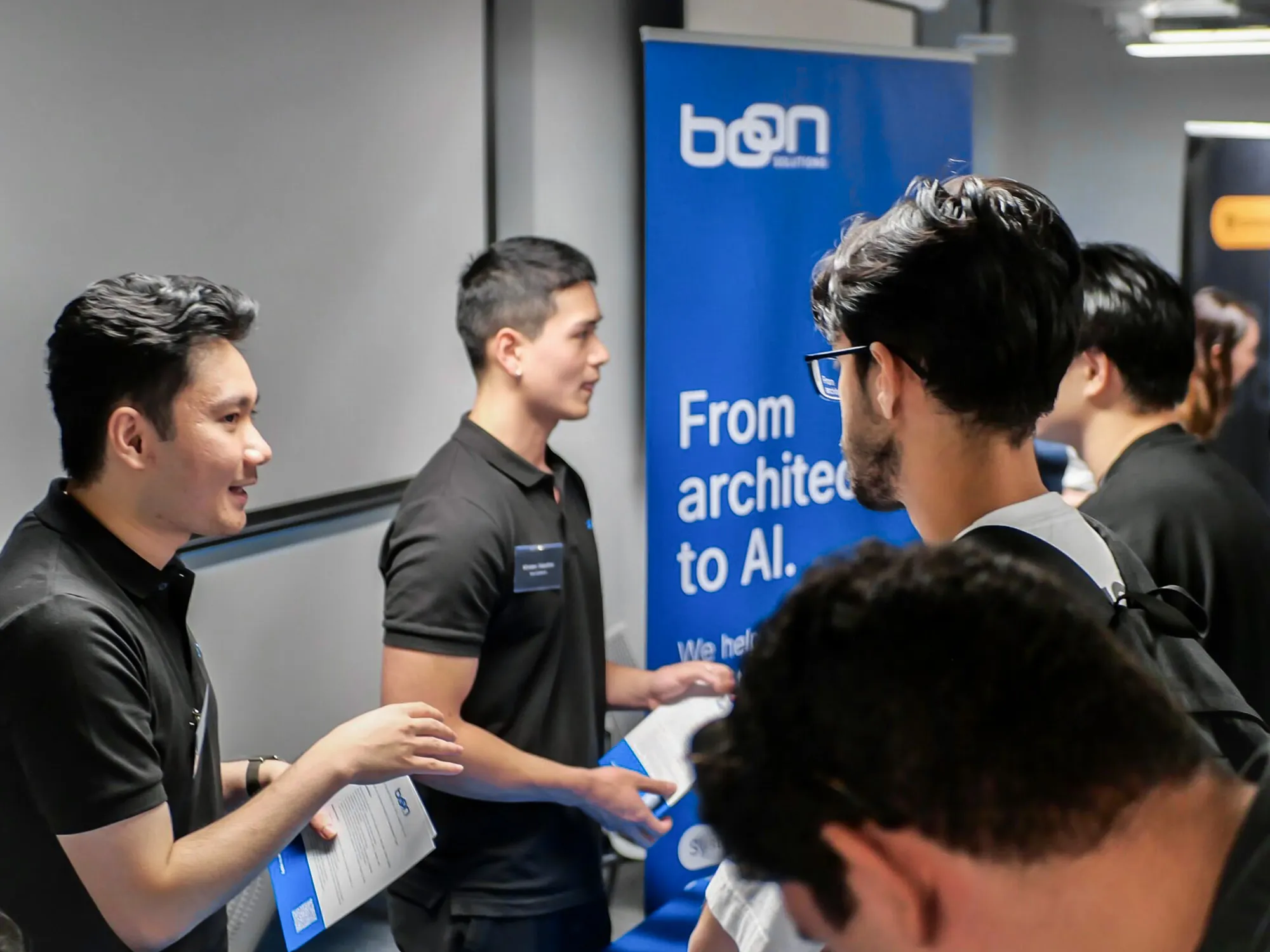Boon Solutions team speaking with attendees beside a blue branded event banner.