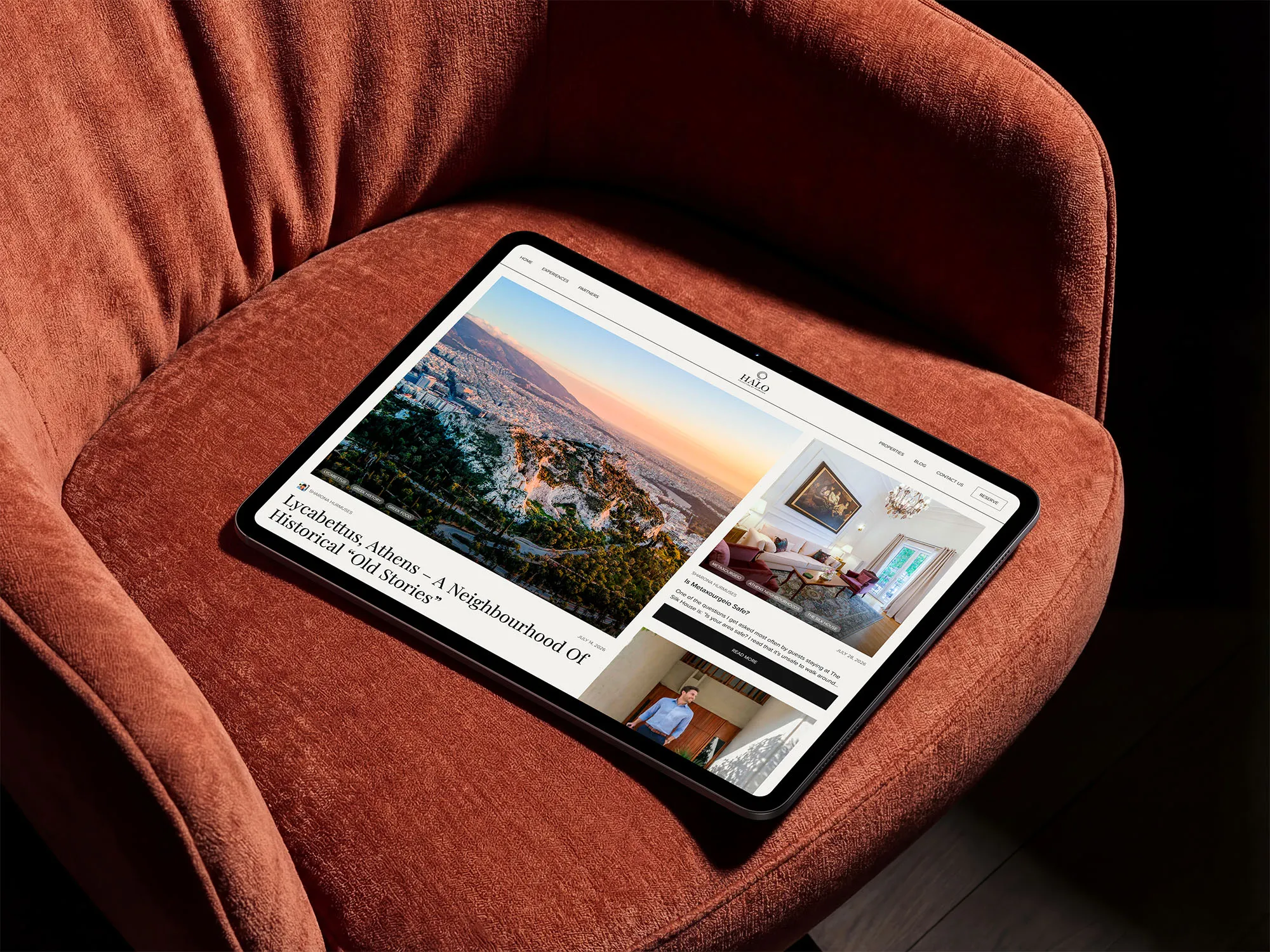 Tablet displaying a website with articles about Athens, including a large photo of Lycabettus Hill at sunset, placed on a textured rust-colored chair.