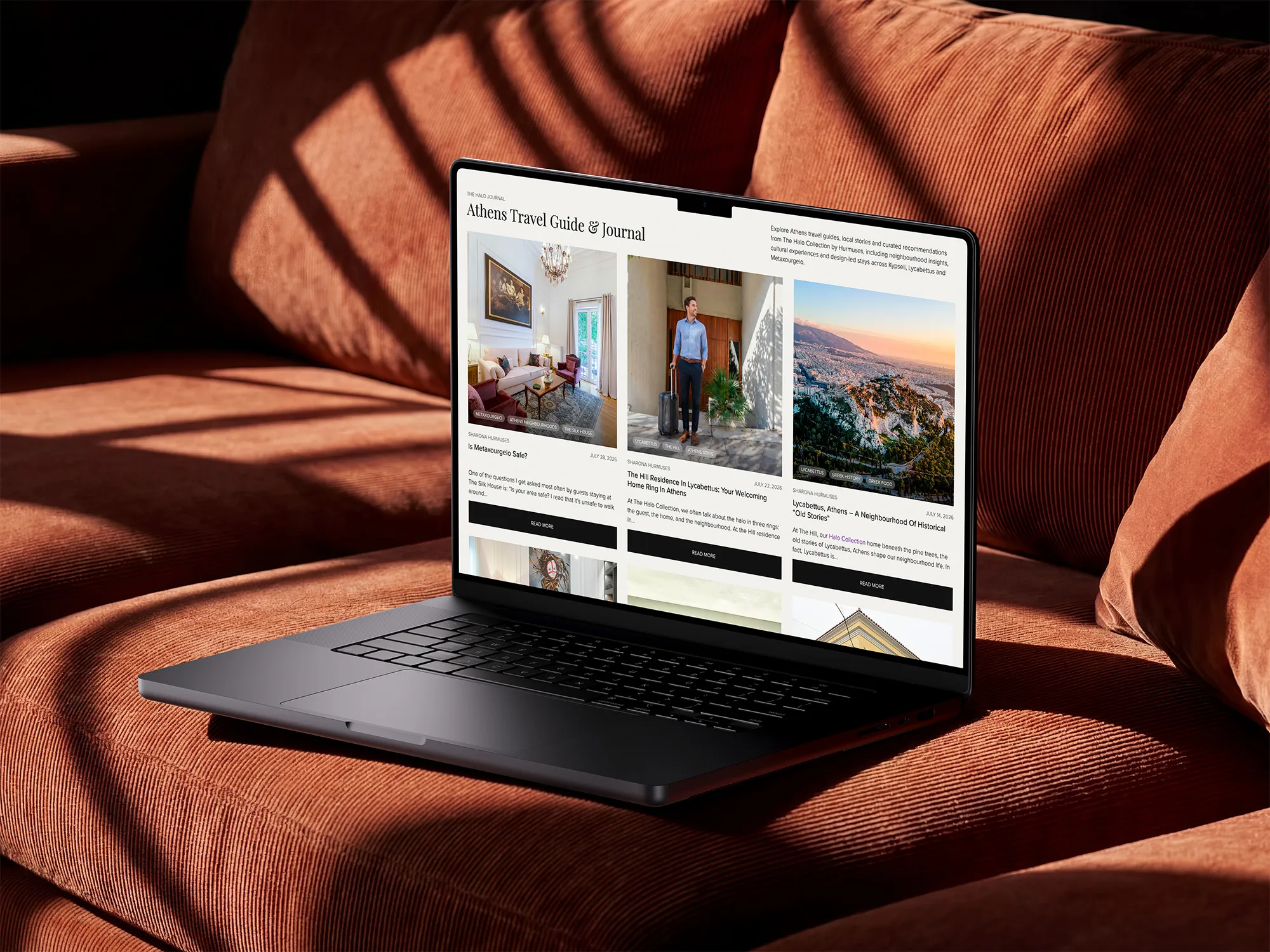 Black laptop on an orange corduroy couch displaying a webpage titled 'Athens Travel Guide & Journal' with travel articles and photos about Athens neighborhoods and culture.