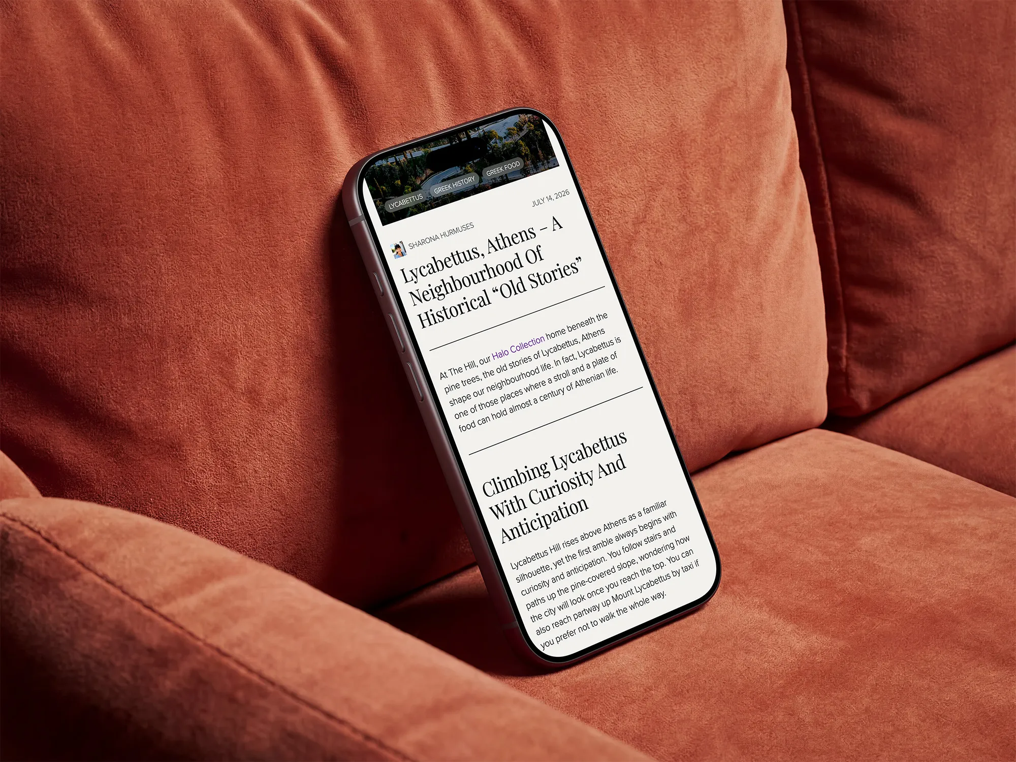 Smartphone resting on a rust-colored fabric couch displaying a blog article titled 'Lycabettus, Athens – A Neighbourhood Of Historical “Old Stories”' by Sharona Hurmuses dated July 14, 2026, with text about Lycabettus Hill and Athenian life.