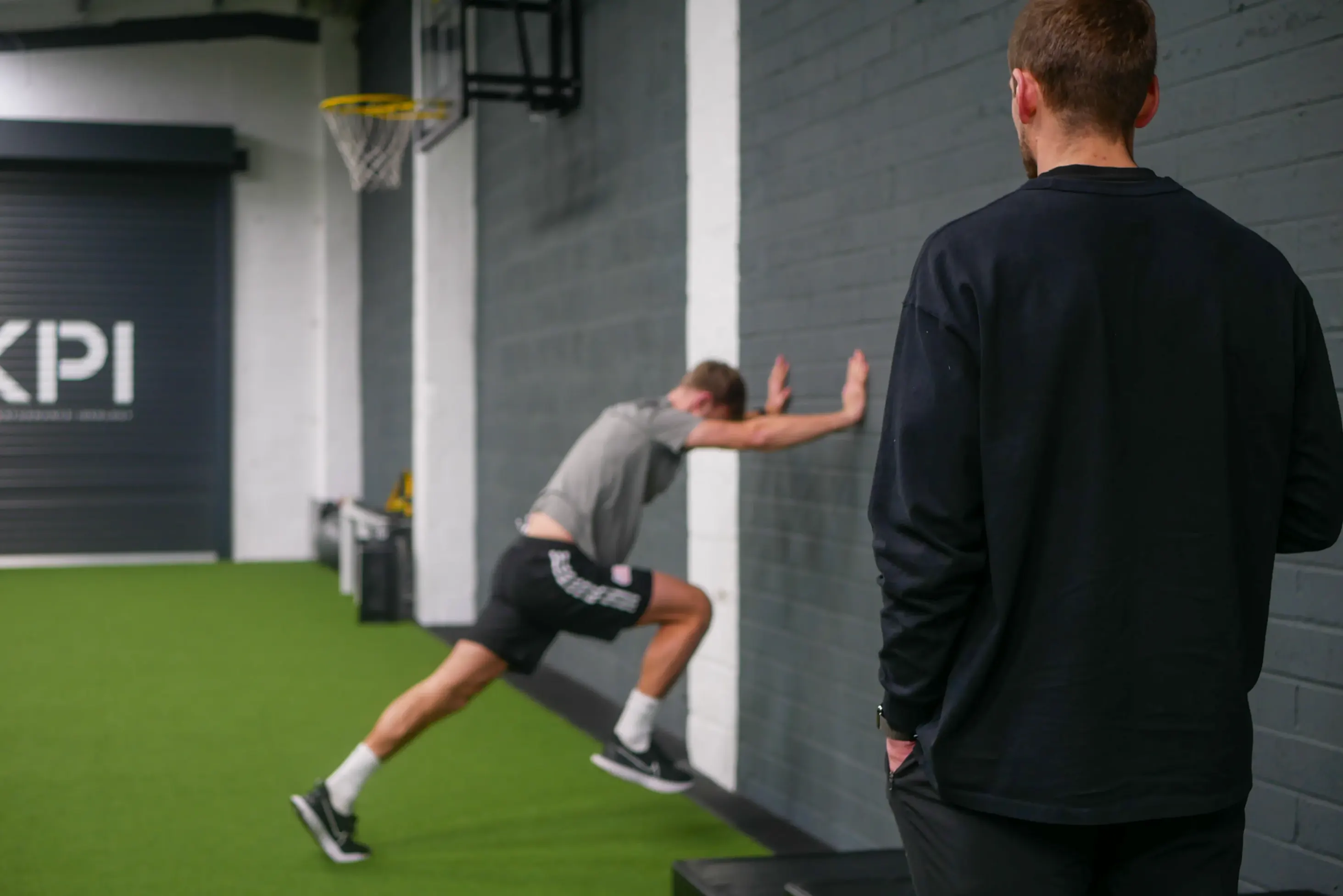 Man in athletic wear stretching against a wall while another man watches in a gym.
