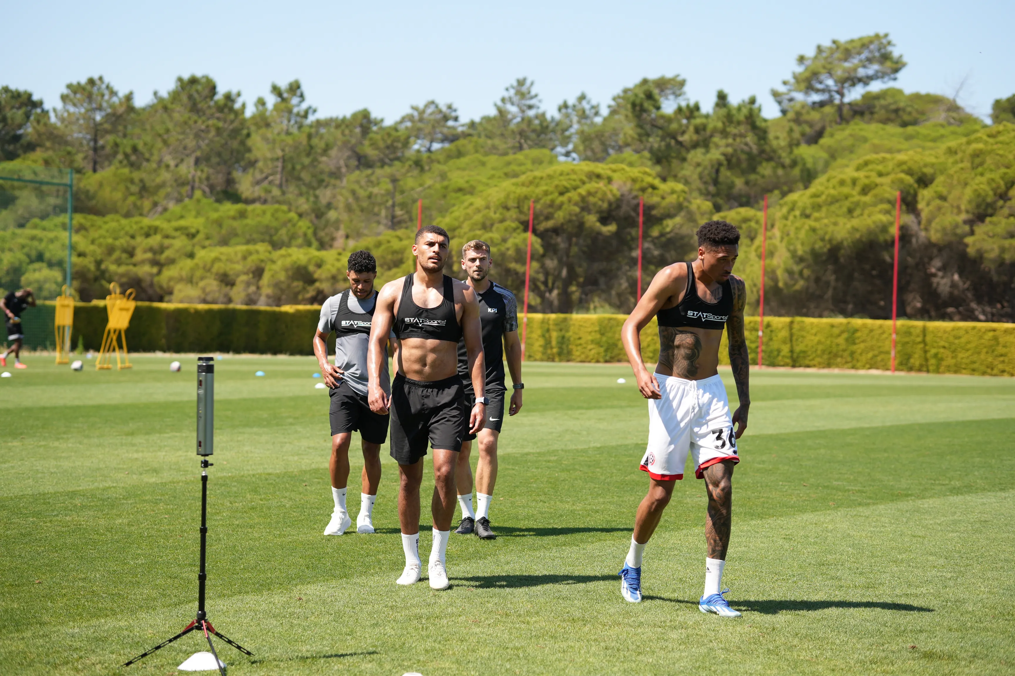 Four male soccer players walking on a green field during daytime training, wearing athletic gear with training vests.