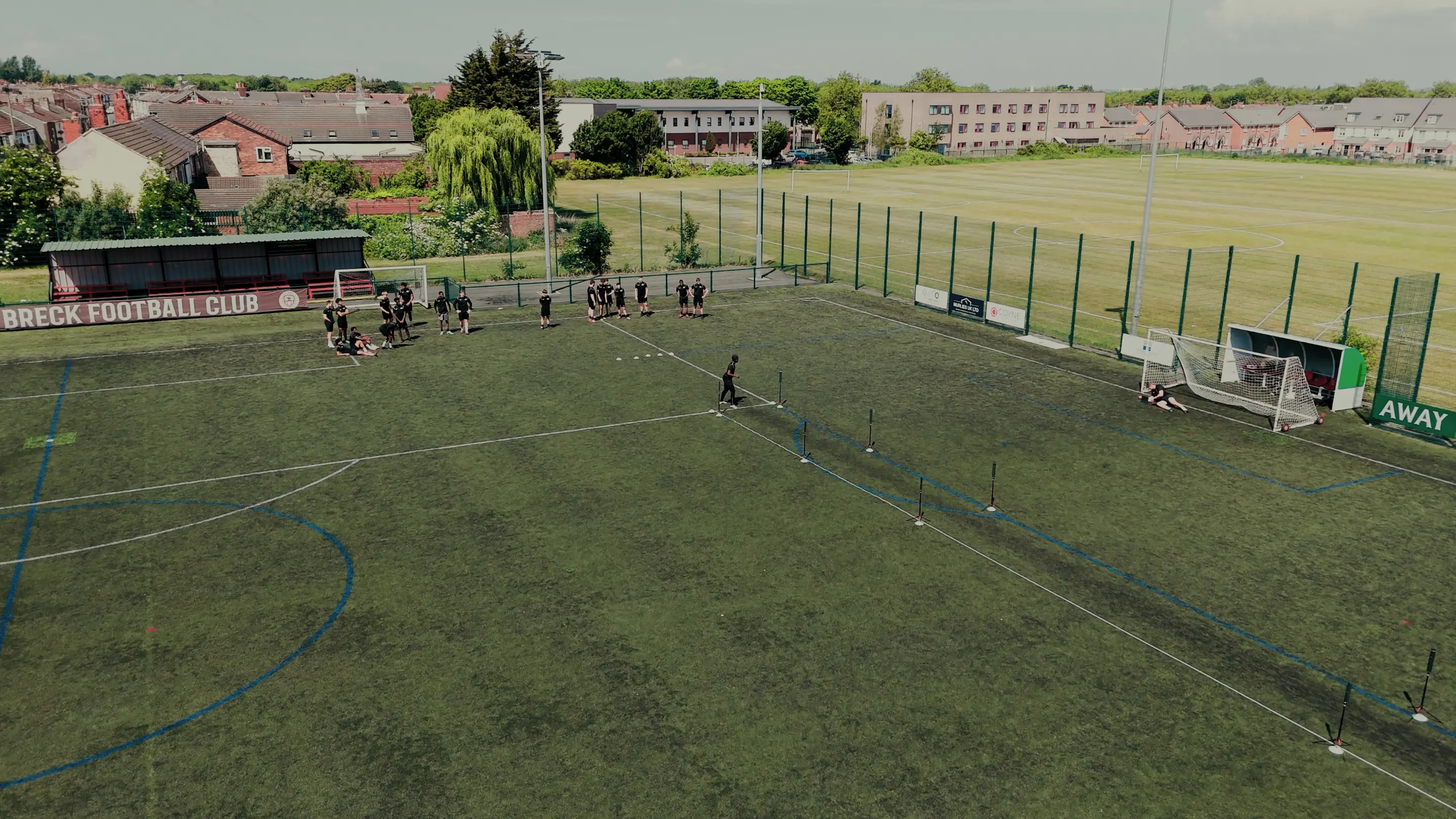 A group of people wearing black on an artificial turf football field near 'Breck Football Club' stand, with a goalkeeper diving near a goal.