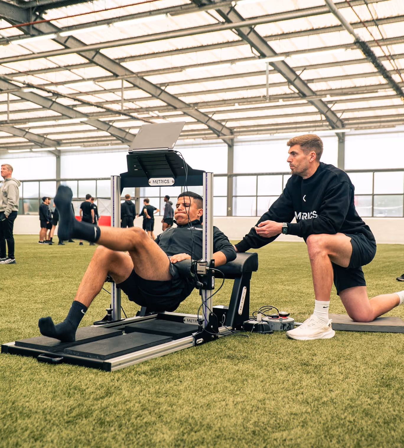 Man performing leg exercise on a METRICS machine while another man kneels beside him, supervising, inside an indoor sports facility.