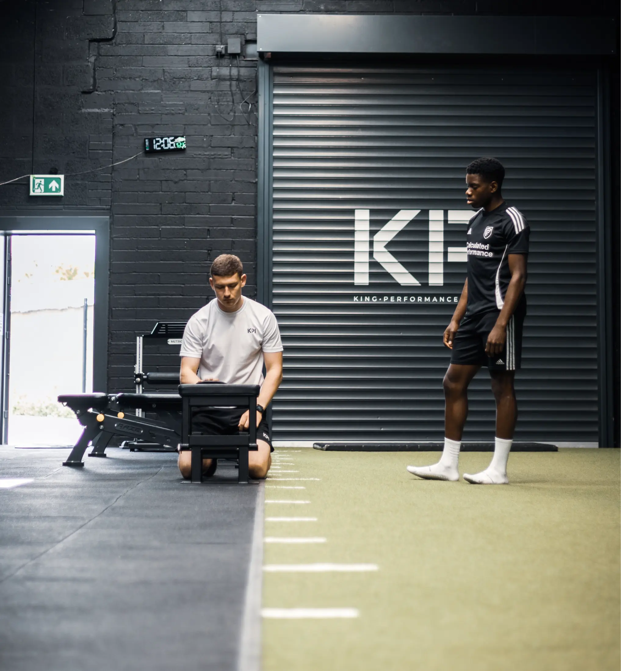 Trainer kneels using device while athlete stands on turf near black wall with large KPI logo.