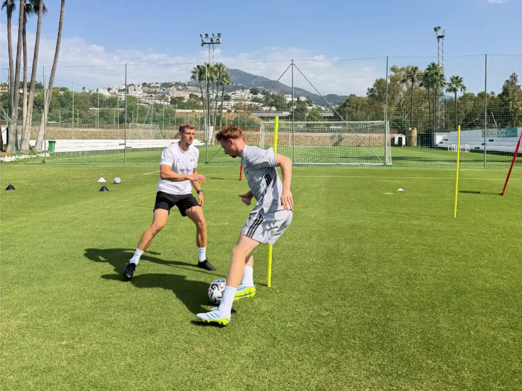 Two men on a grass field practicing soccer dribbling around yellow poles and cones under blue sky.