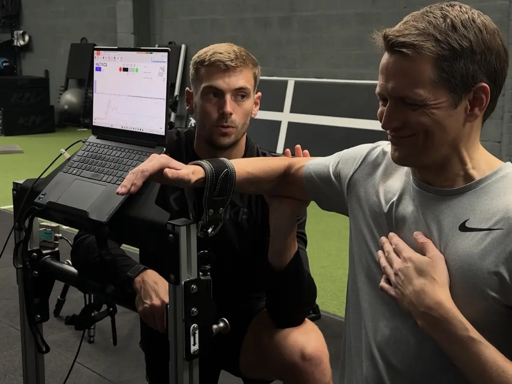 Trainer assisting man doing arm strength exercise with a device and laptop displaying metrics.