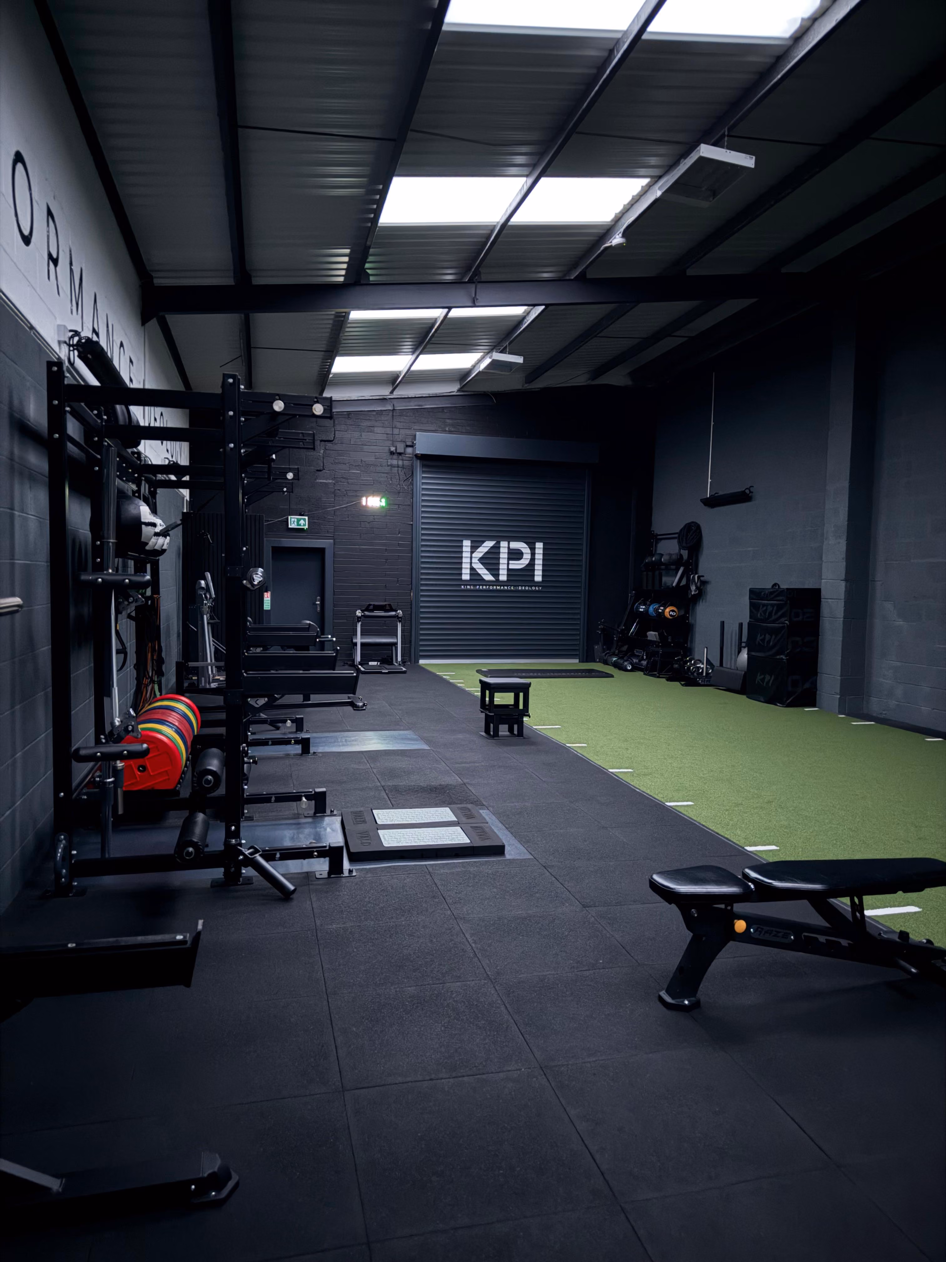 Indoor gym with black flooring and green turf area featuring weightlifting racks with colorful weight plates, a workout bench, a small plyometric box, and various fitness equipment along dark walls under a metal roof with skylights.