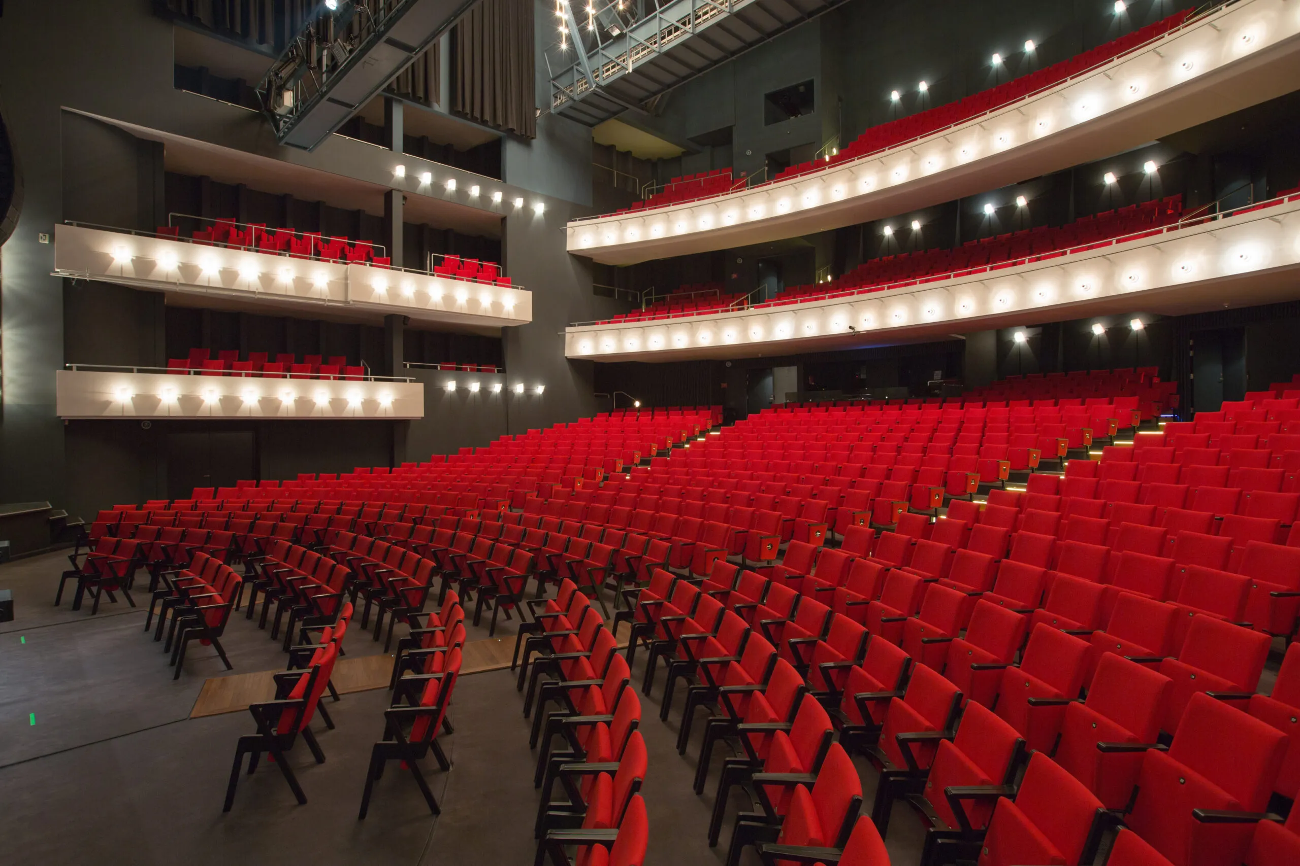 Interior of a large modern theater with rows of red seats on the main floor and two balconies above, all facing the stage area, illuminated by warm lighting.