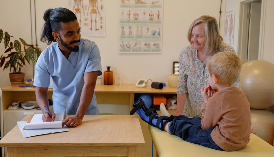 A male healthcare worker in blue scrubs taking notes in an examination room next to a young boy sitting on an examining table next to his mom.