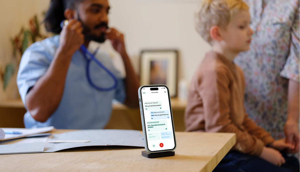 A male healthcare worker in blue scrubs is examining a boy with a sthetoscope as the boys mother is standing next to him. In the foreground you can see a smartphone with the interface of the Care to Translate translation app.