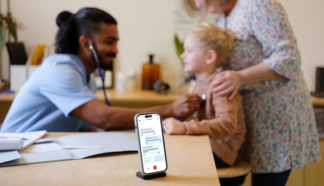 A male healthcare professional in blue scrubs examining a young boy with a stethoscope while the boys mother stands behind the boy. A smartphone with the Care to Translate app in hands-free mode can be seen in the foreground.