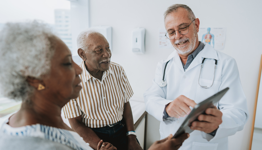 male doctor in white coat showin two elderly patients something on a tablet in a light examination room