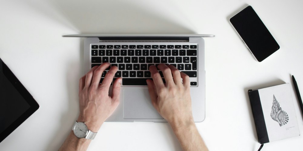 Hands typing on a laptop on a white desk beside a phone and a small notebook