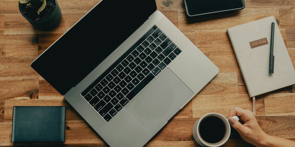 Laptop, notebook, phone, and a cup of coffee on a wooden desk seen from above