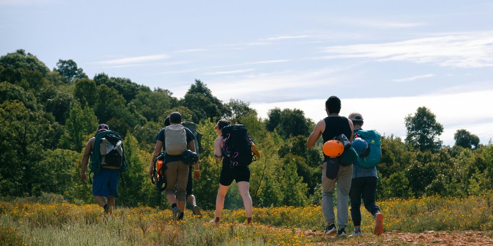 Group of hikers carrying backpacks and climbing gear walking together across a sunny meadow toward a tree line
