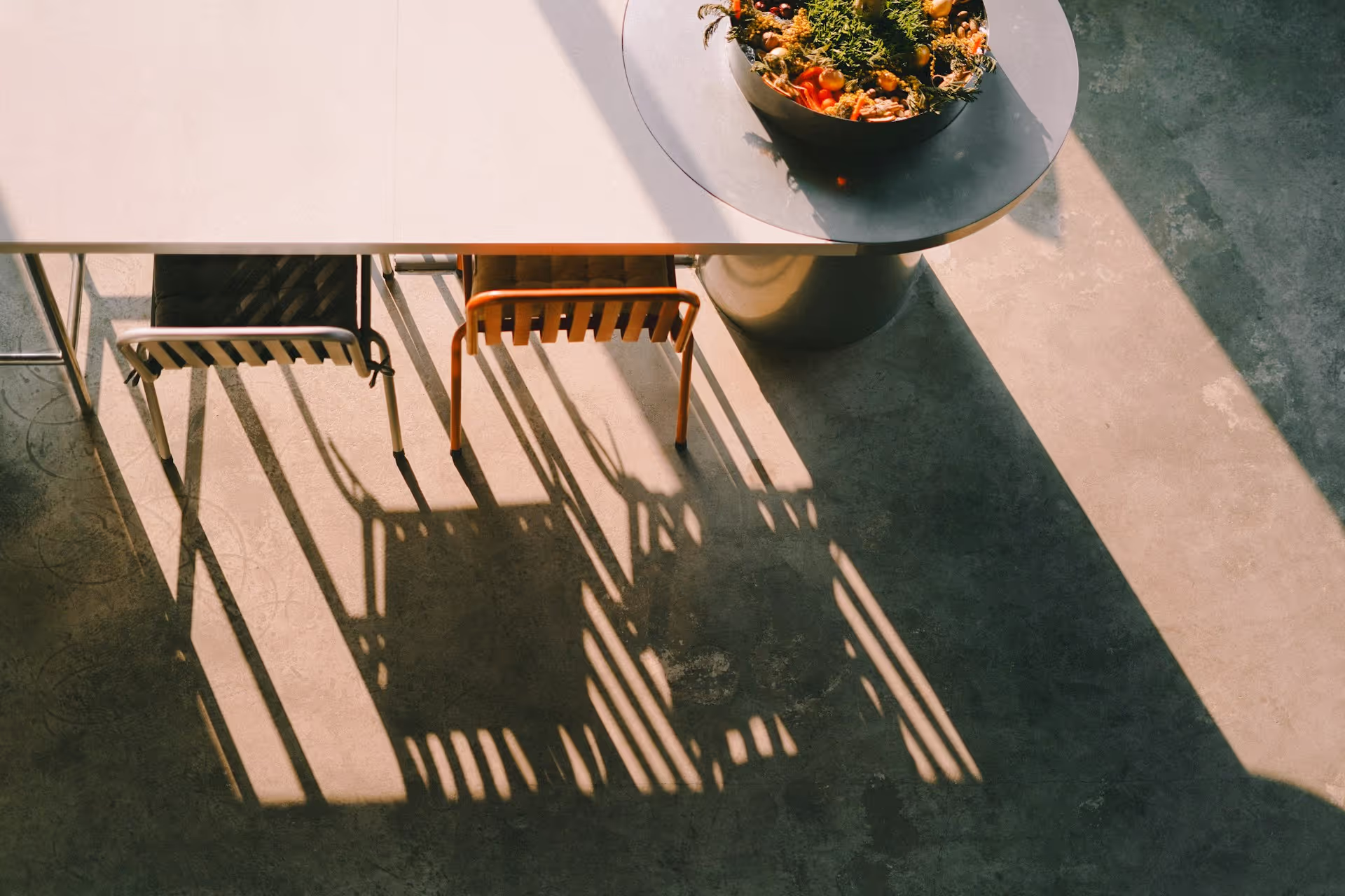 Two chairs casting long shadows on a concrete floor beside a white table and a round side table holding a decorative plant bowl.