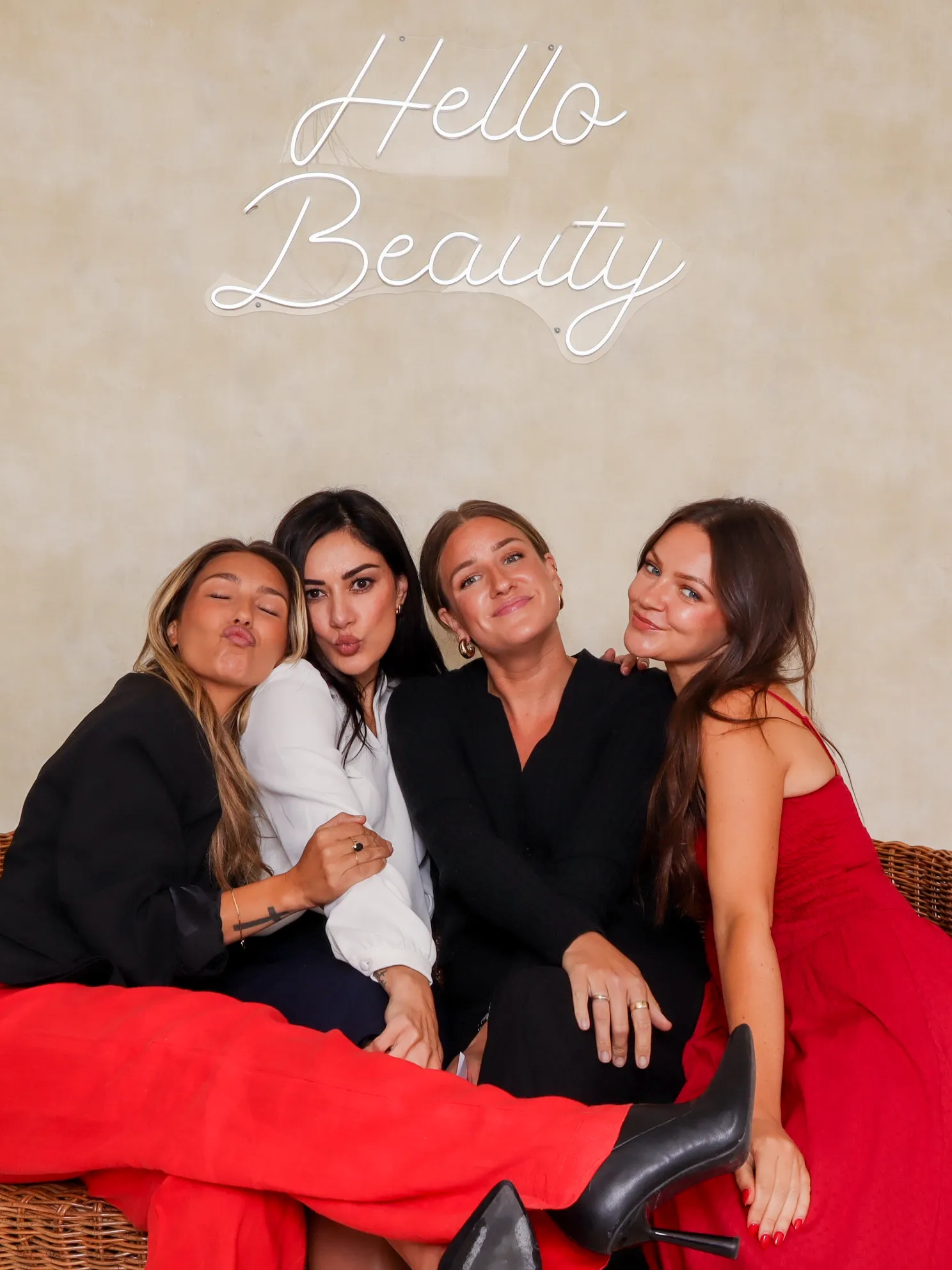 Four women sitting closely together on a wicker sofa, smiling and posing under a neon sign that says 'Hello Beauty'.