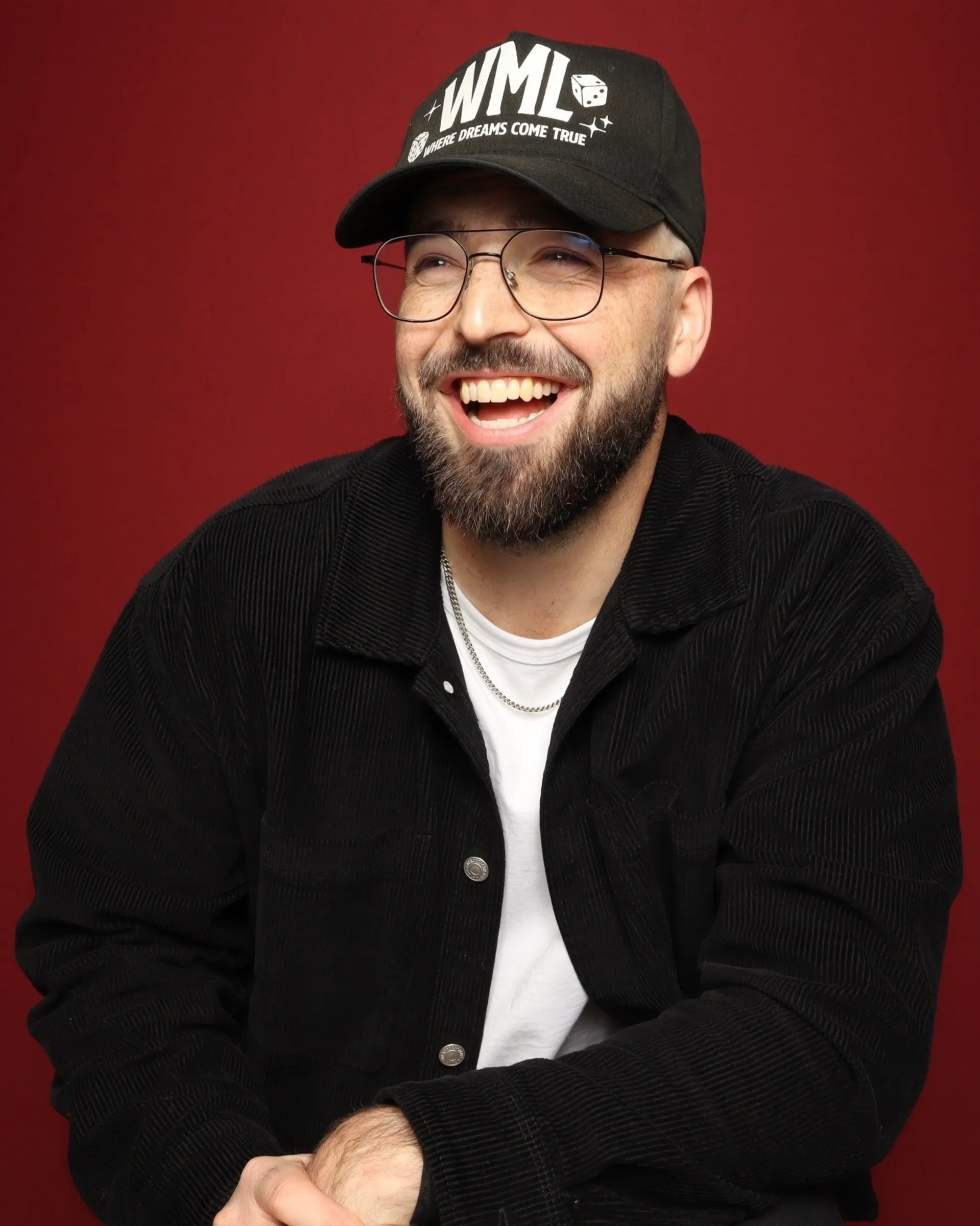Portland portrait photo booth rental. Smiling man wearing glasses, a black cap, black jacket, white shirt, and gold chain against a red background.