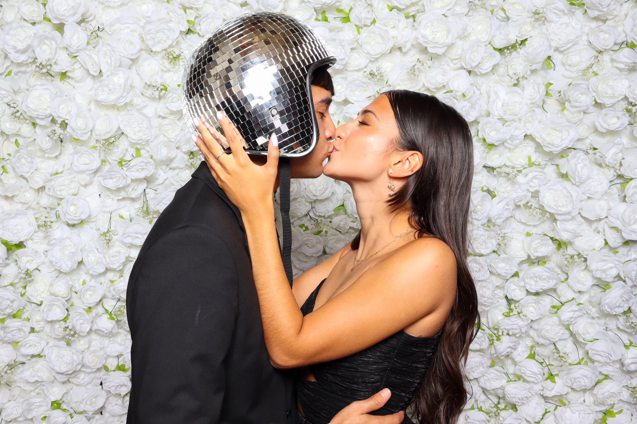 Couple kissing in front of a white rose flower wall, the man wears a disco ball helmet and a black suit, the woman wears a black dress.