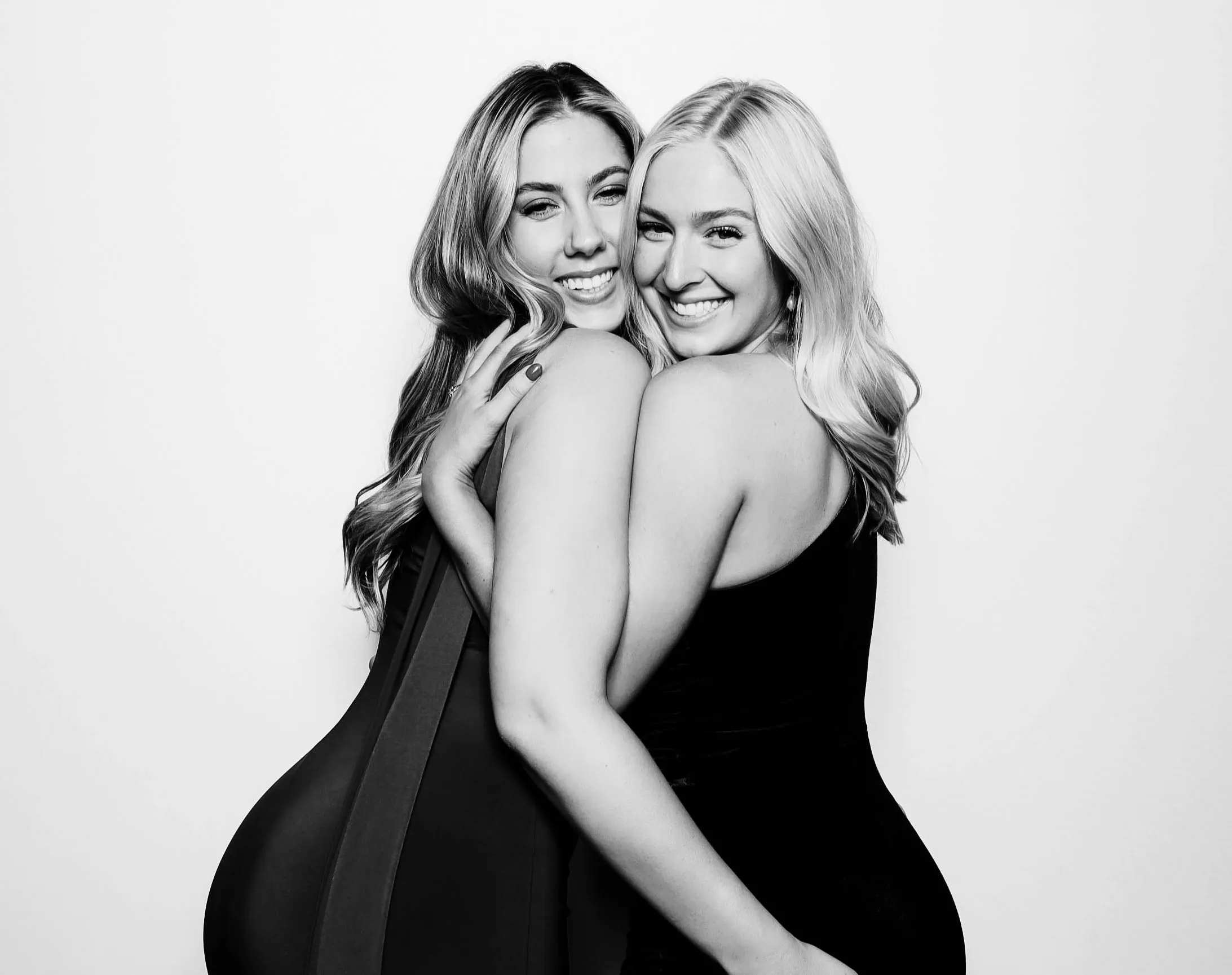 Two smiling women with long hair embracing and posing against a plain background in a black and white glam photo booth photo.