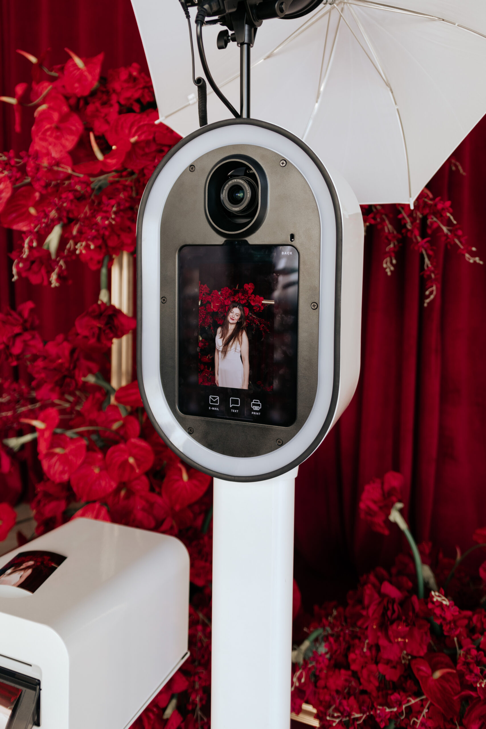 Photo booth kiosk with digital screen displaying a woman posing in front of a red floral backdrop.