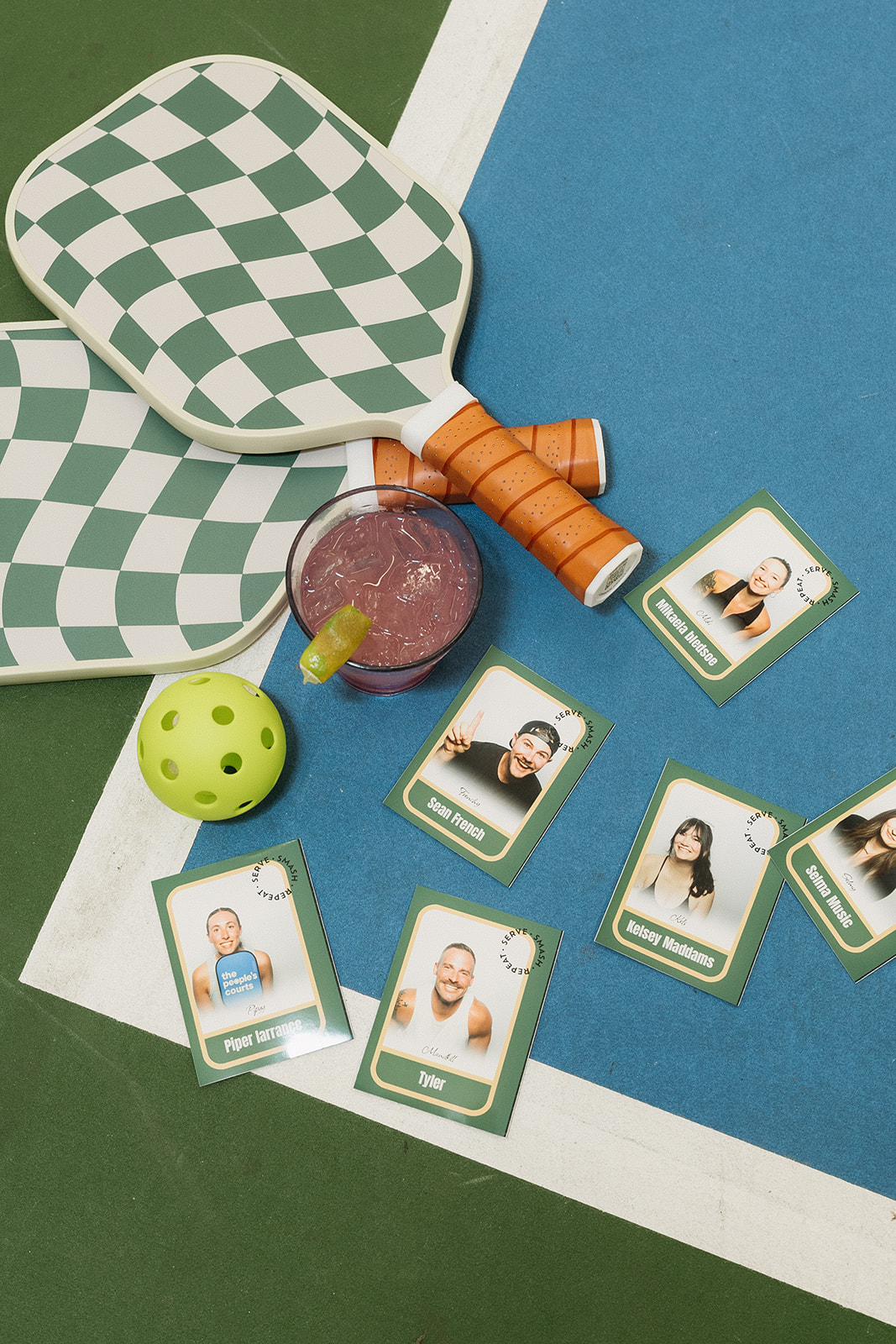 Two green and white checkered pickleball paddles, a yellow pickleball, a glass with a pink drink and lime wedge, and six custom photo booth printed player cards with photos on a blue and green court surface.
