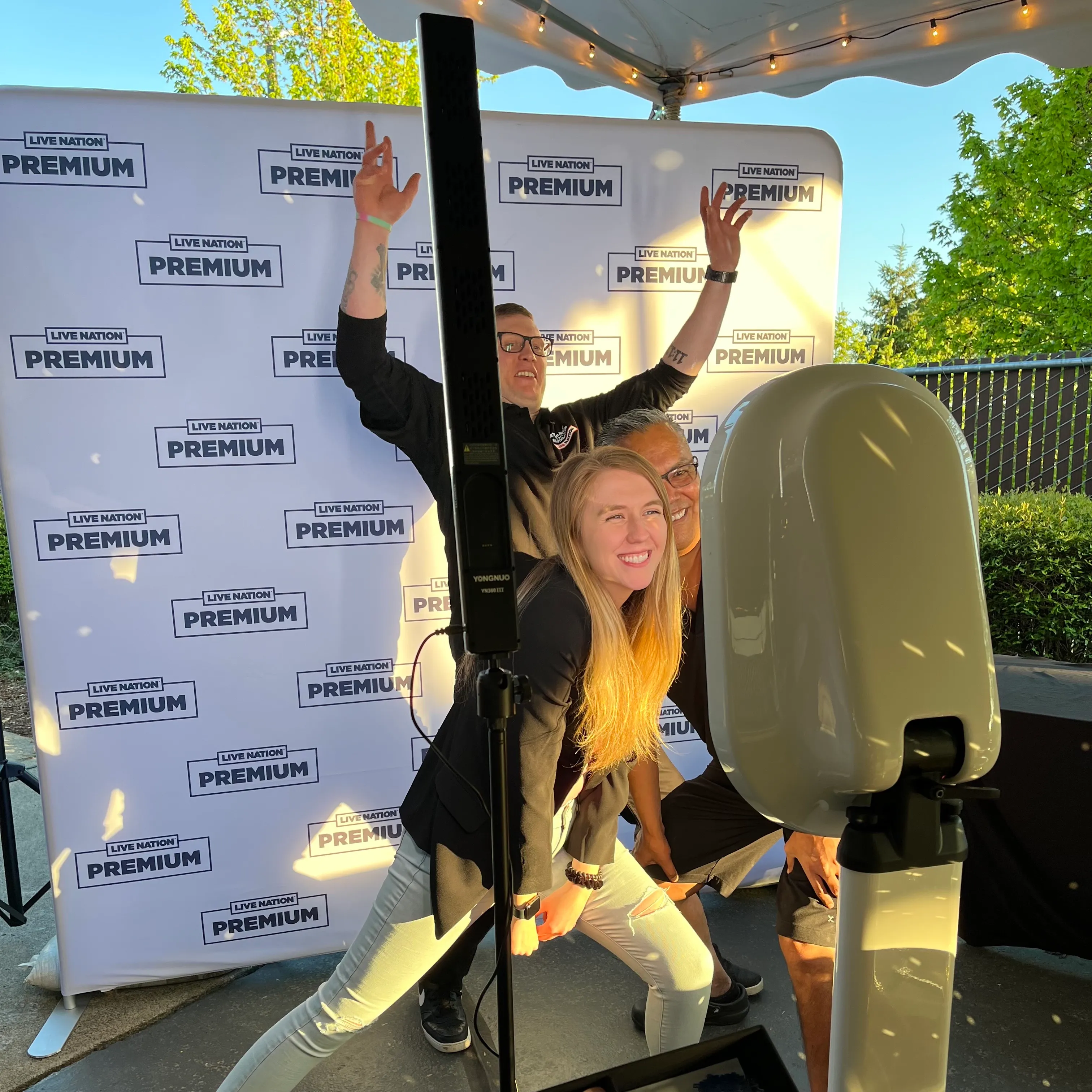 Three people posing and smiling in front of a Live Nation Premium backdrop at a photo booth under a tent.