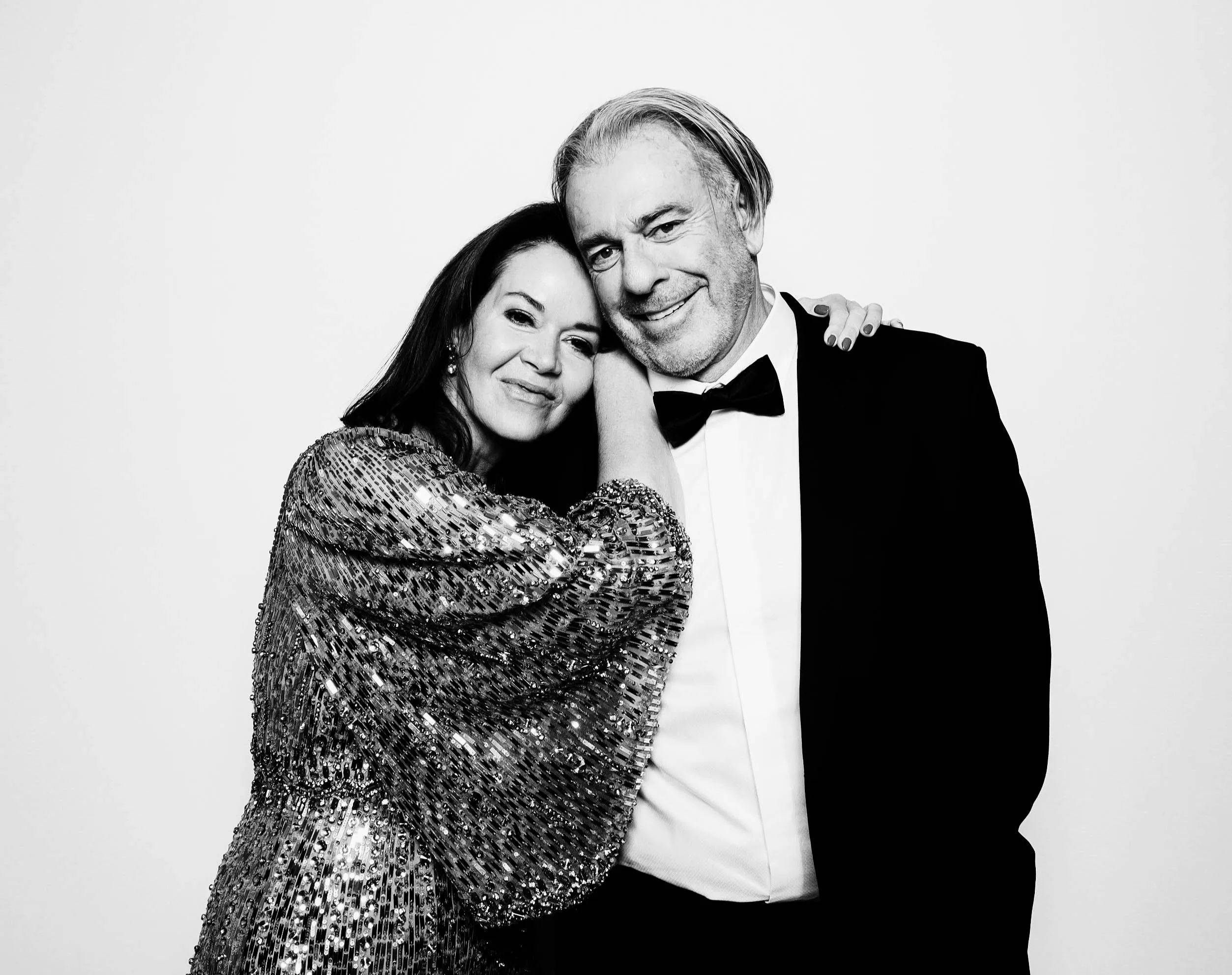 Smiling couple posing closely in a glam photo booth in Portland, the woman wearing a sequined dress with her arm around the man's shoulder, who is dressed in a tuxedo with a bow tie.
