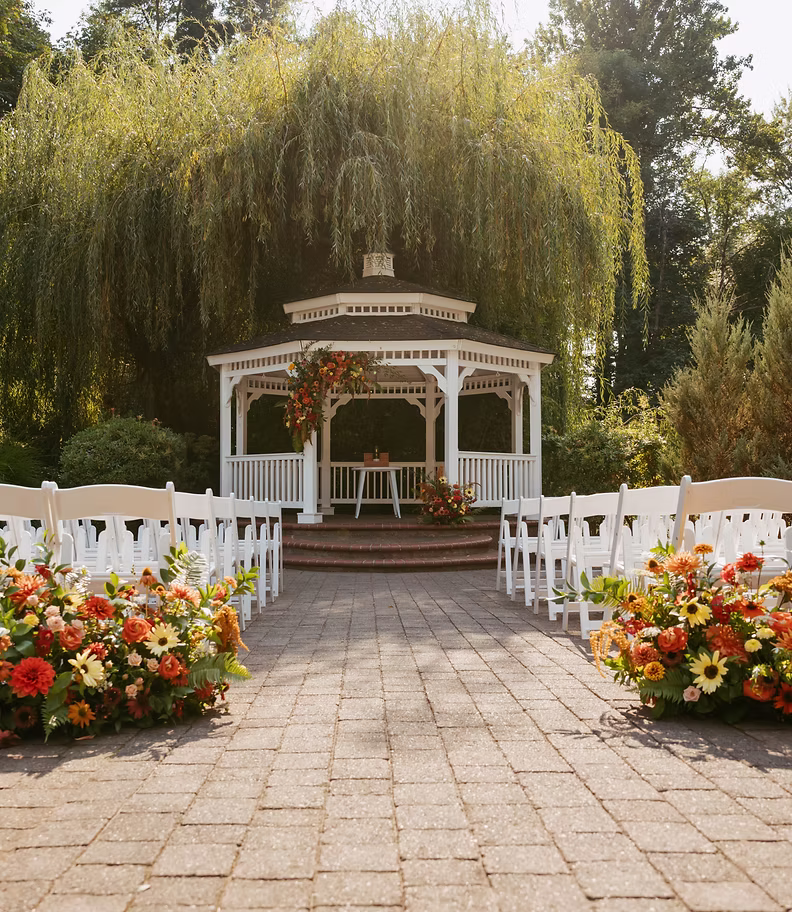 Outdoor wedding setup at Abernethy Center with a white gazebo decorated with floral arrangements and rows of white chairs on both sides of a stone pathway.