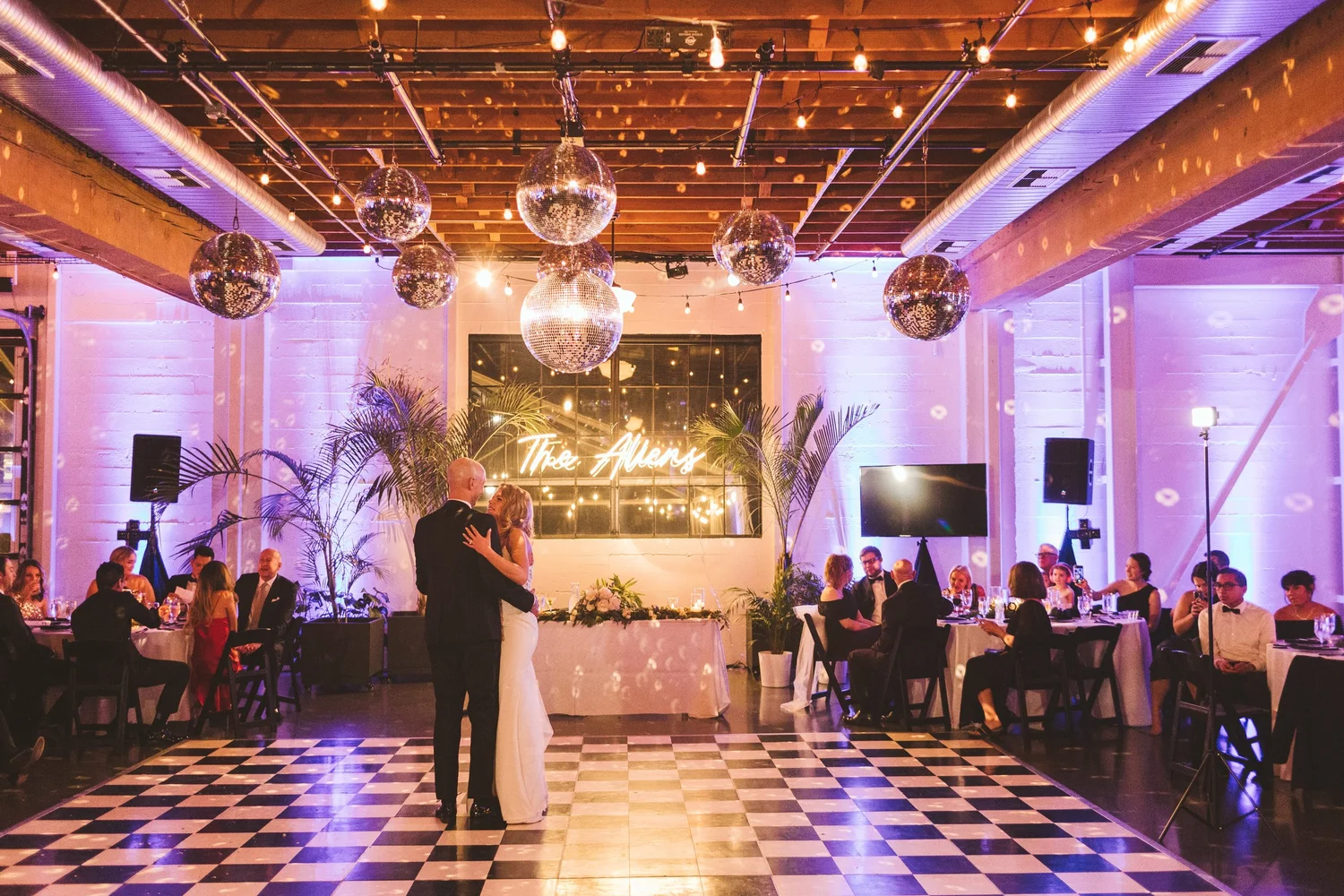 Bride and groom dancing on a black and white checkered floor at The Castaway in Portland with guests seated at round tables under hanging disco balls and string lights.