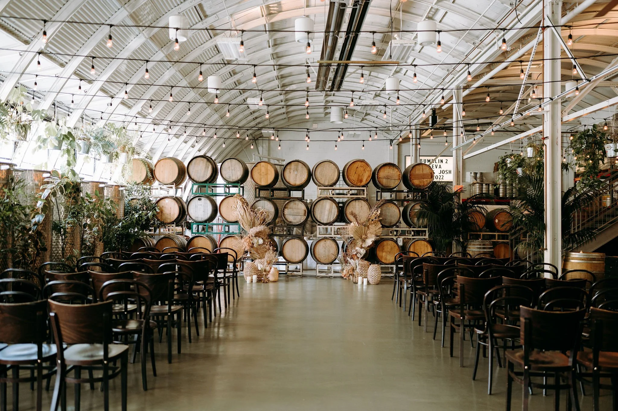 Indoor event space at Coopers Hall with rows of wooden chairs facing stacked wooden barrels and dried floral arrangements under string lights.