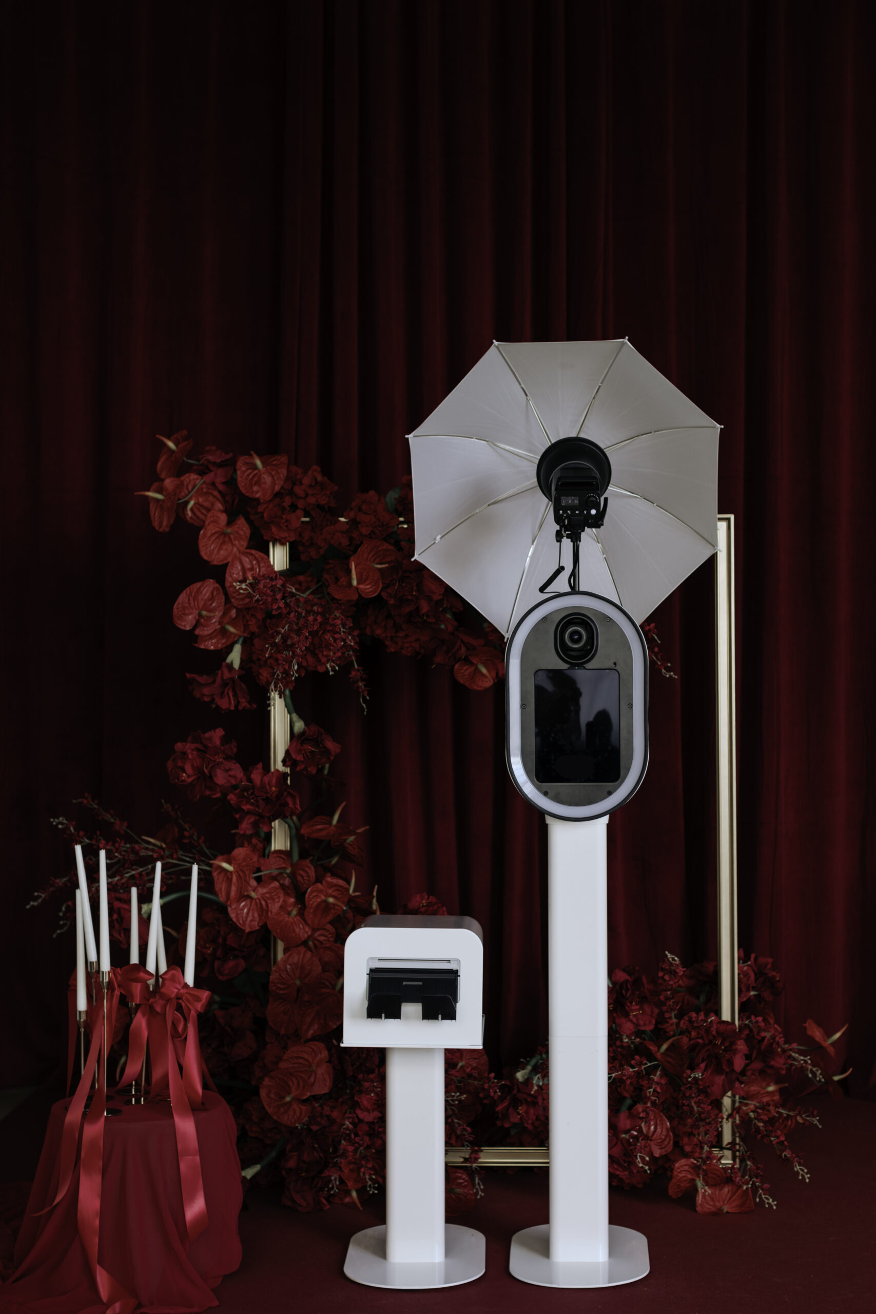 Photo booth setup with a camera and white umbrella light on stands, surrounded by red flowers and candles with red ribbons in front of a red curtain.