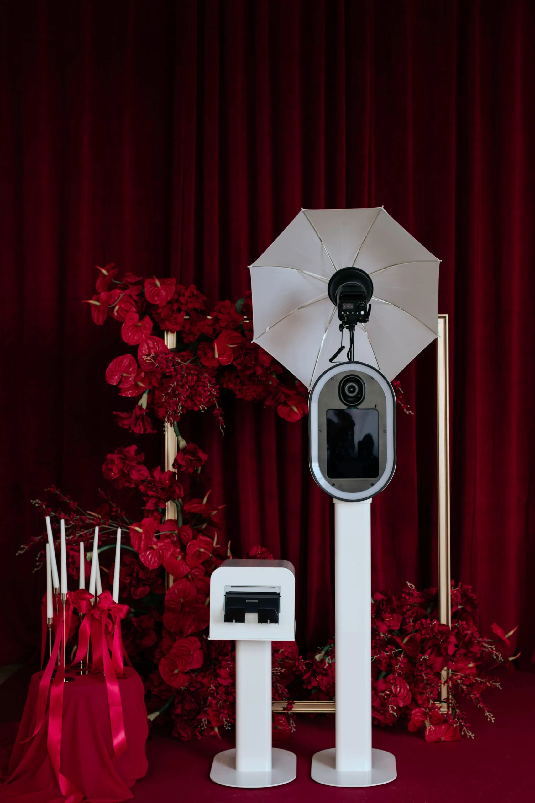 Photo booth setup with a camera and white umbrella light on stands, surrounded by red flowers and candles with red ribbons in front of a red curtain.