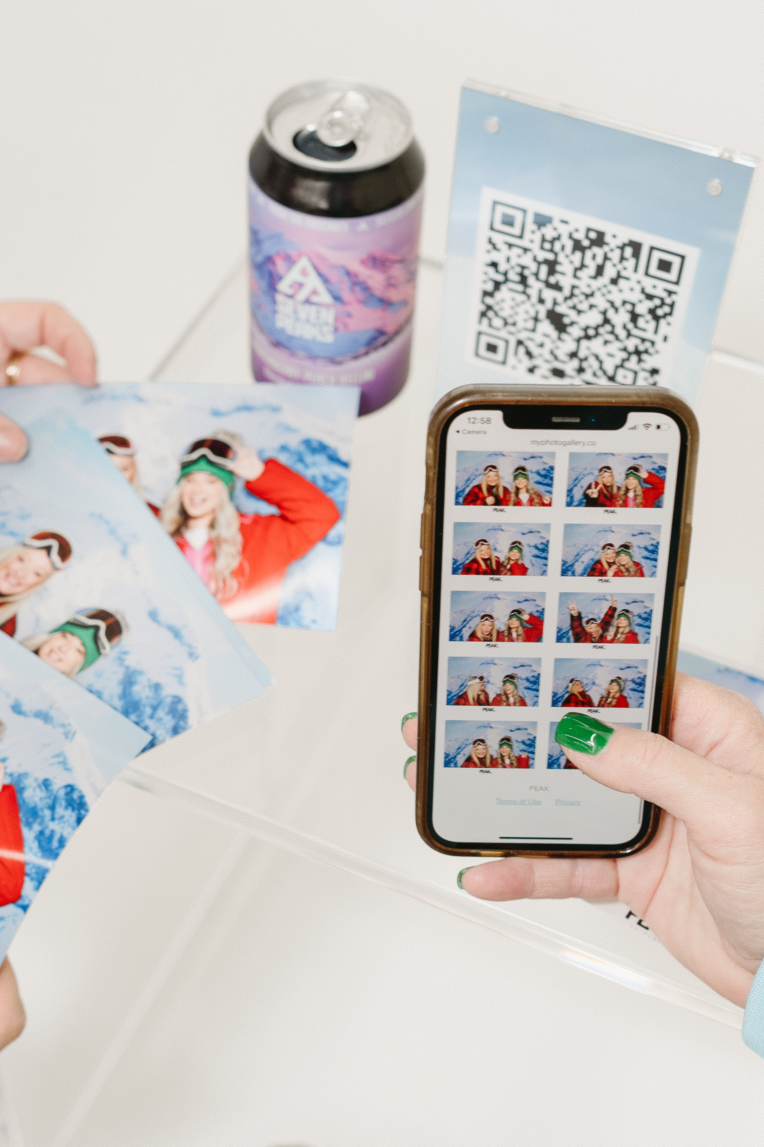 Close-up of a hand holding a smartphone displaying photo booth images of two women in winter gear next to printed photos and a drink can with a QR code sign on a clear table.