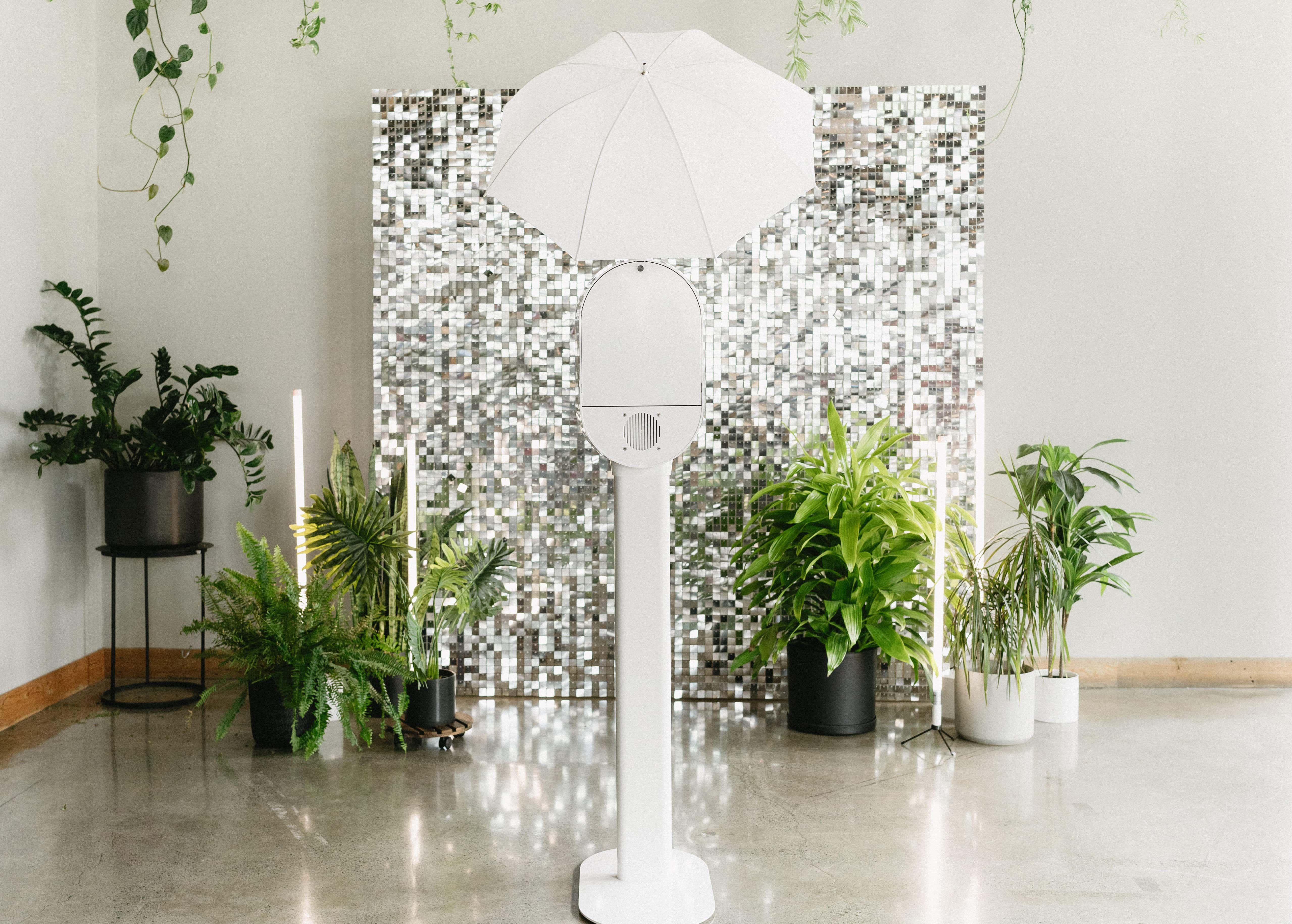 White photo booth with umbrella light in front of a reflective mosaic backdrop, surrounded by green potted plants on a polished concrete floor.