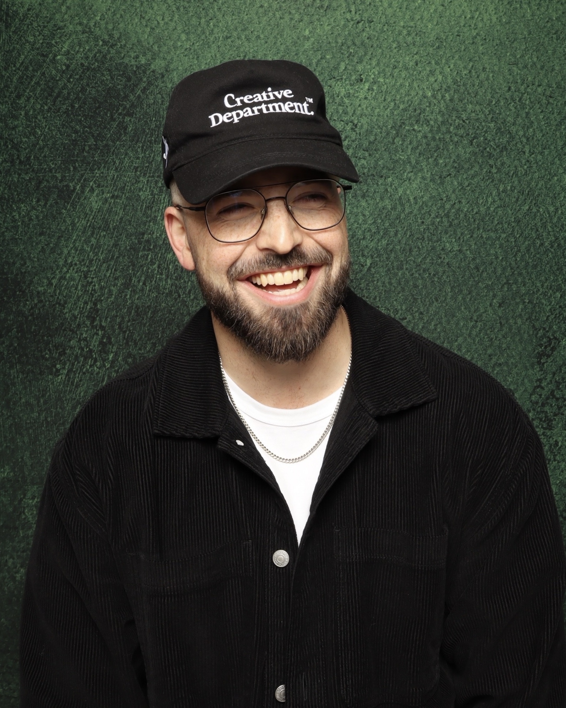 Portrait photo booth photo of a smiling bearded man wearing glasses, a black 'Creative Department' cap, black jacket, and a gold chain necklace in front of a green textured background.