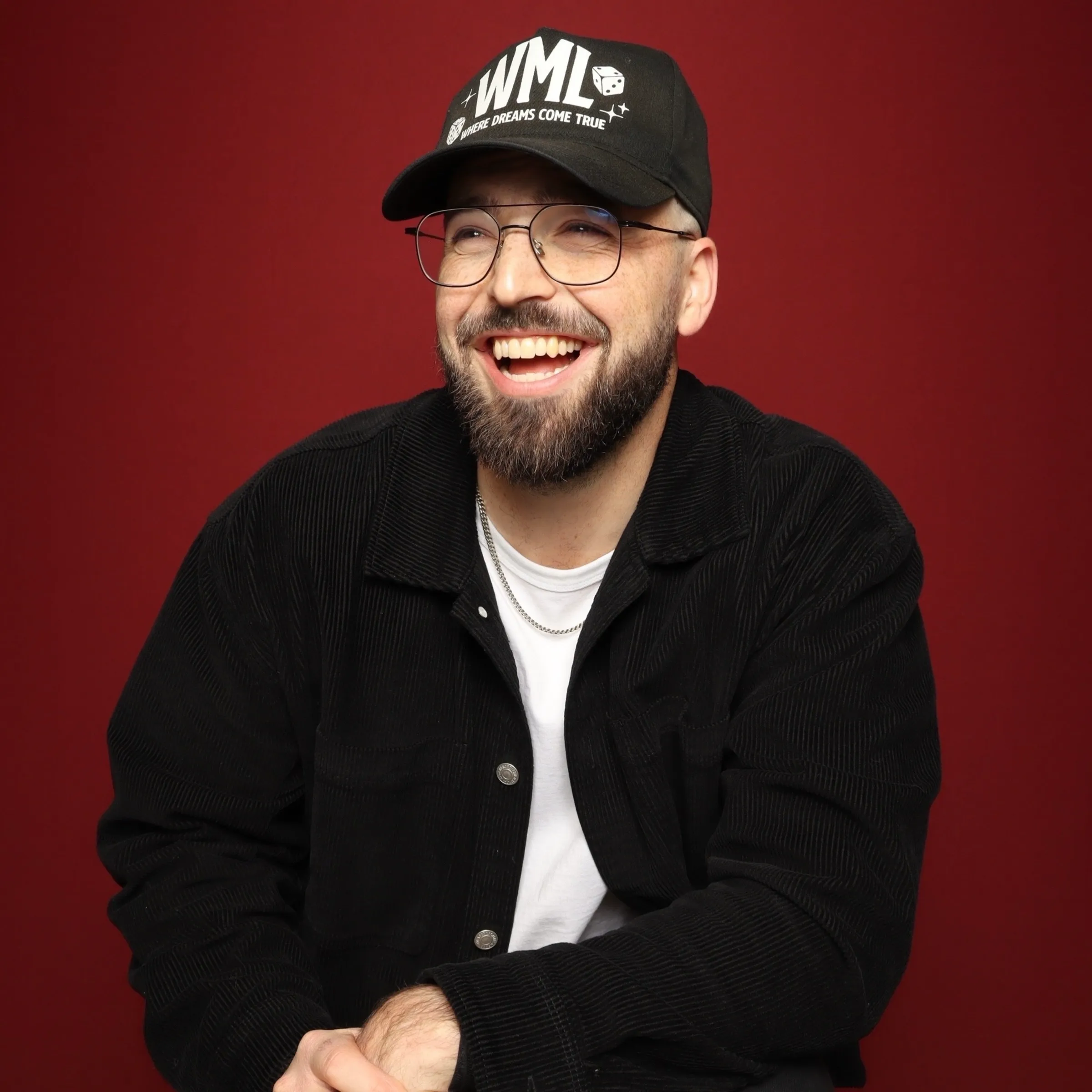 Smiling man with beard and glasses wearing a black cap and jacket against a red background.